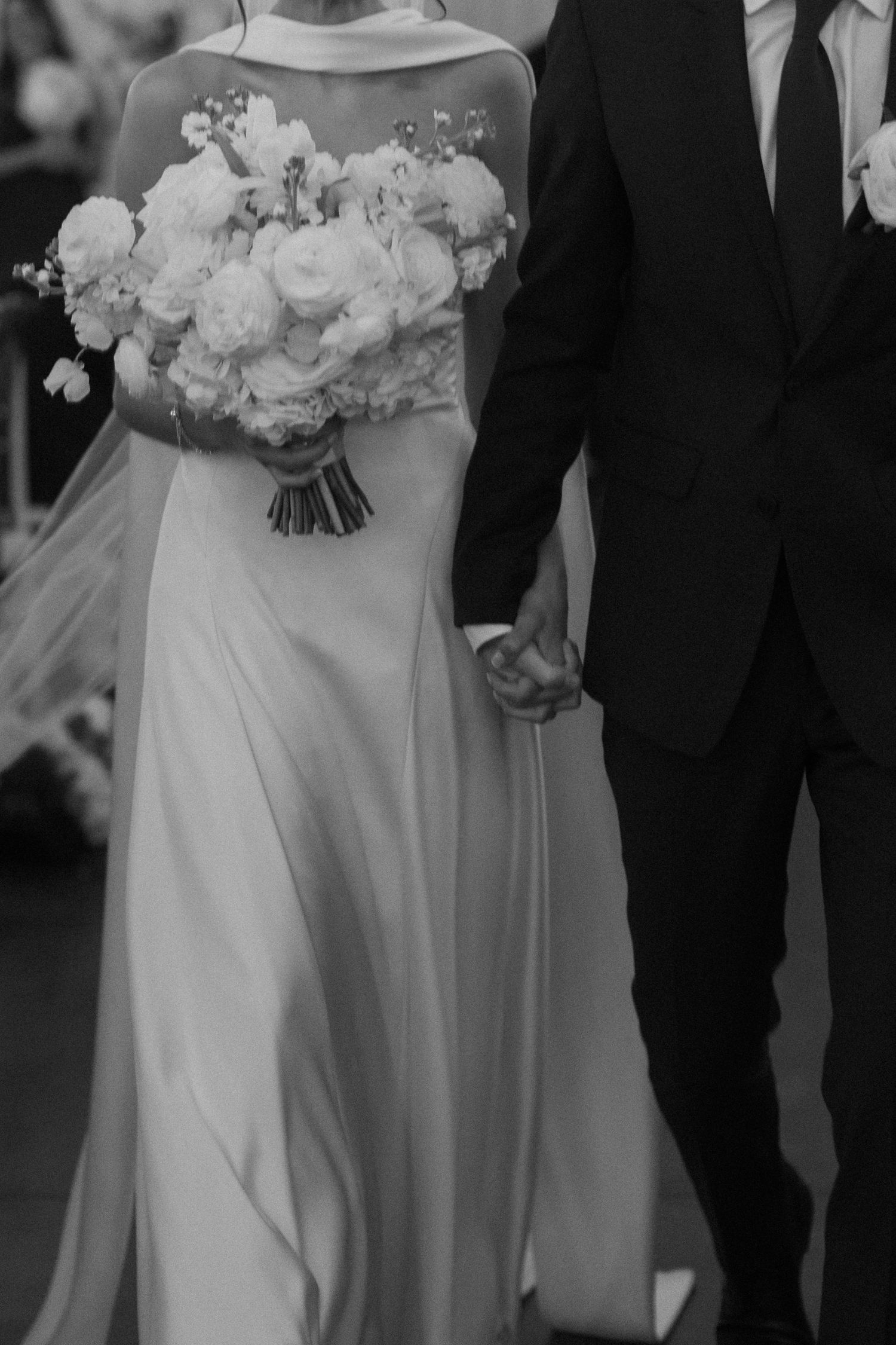 Black and white wedding photo showing elegant bride in flowing dress holding white bouquet beside groom in suit.