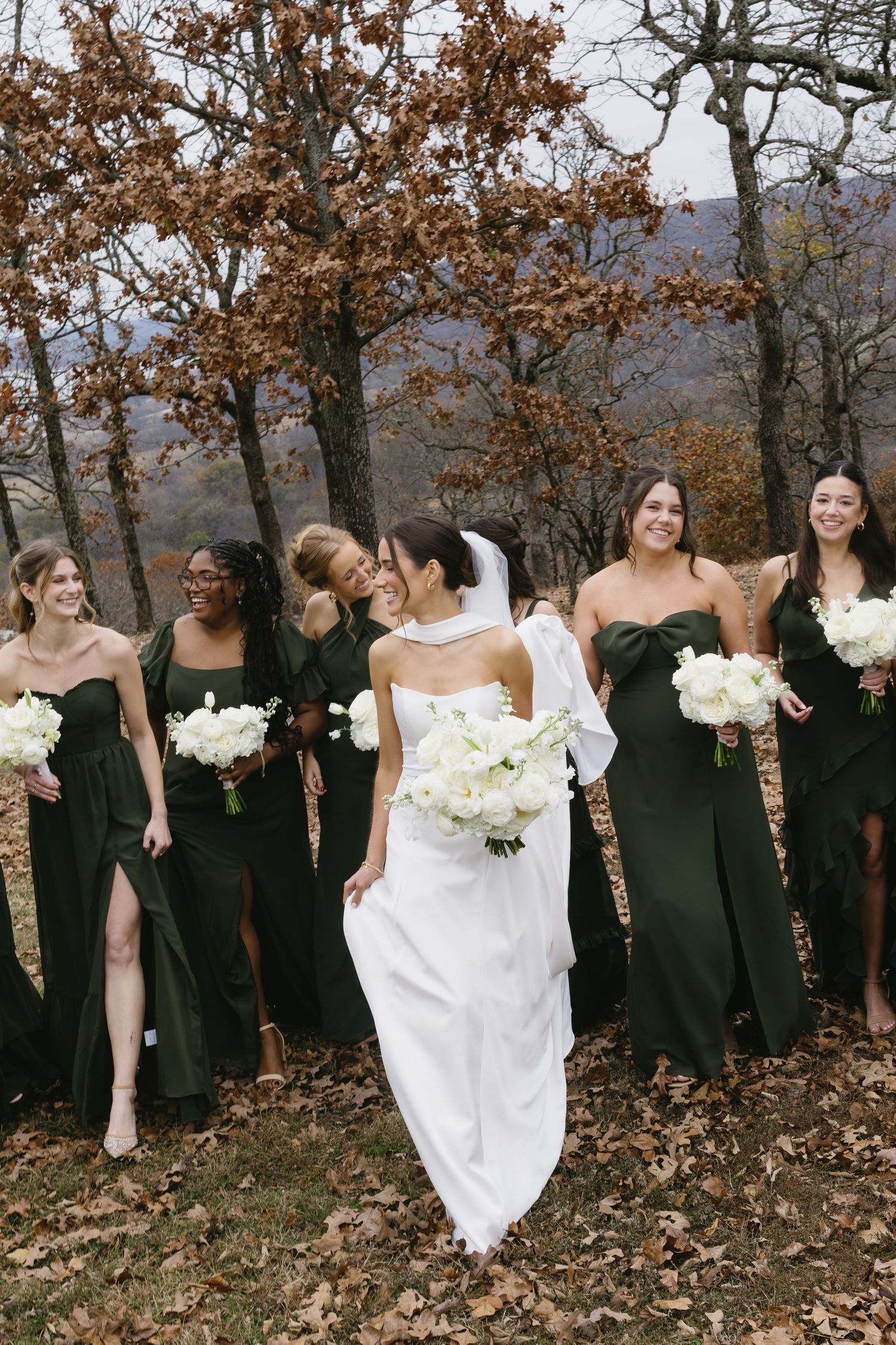 Wedding party poses in autumn setting with bridesmaids in black dresses and bride in white holding bouquets.