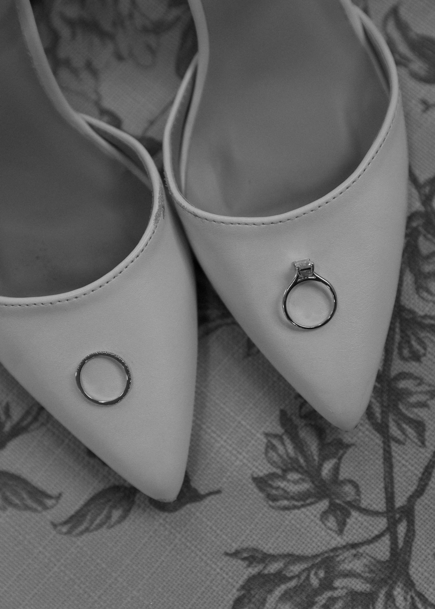 Close-up of elegant white bridal shoes with engagement rings placed on top against a floral background.