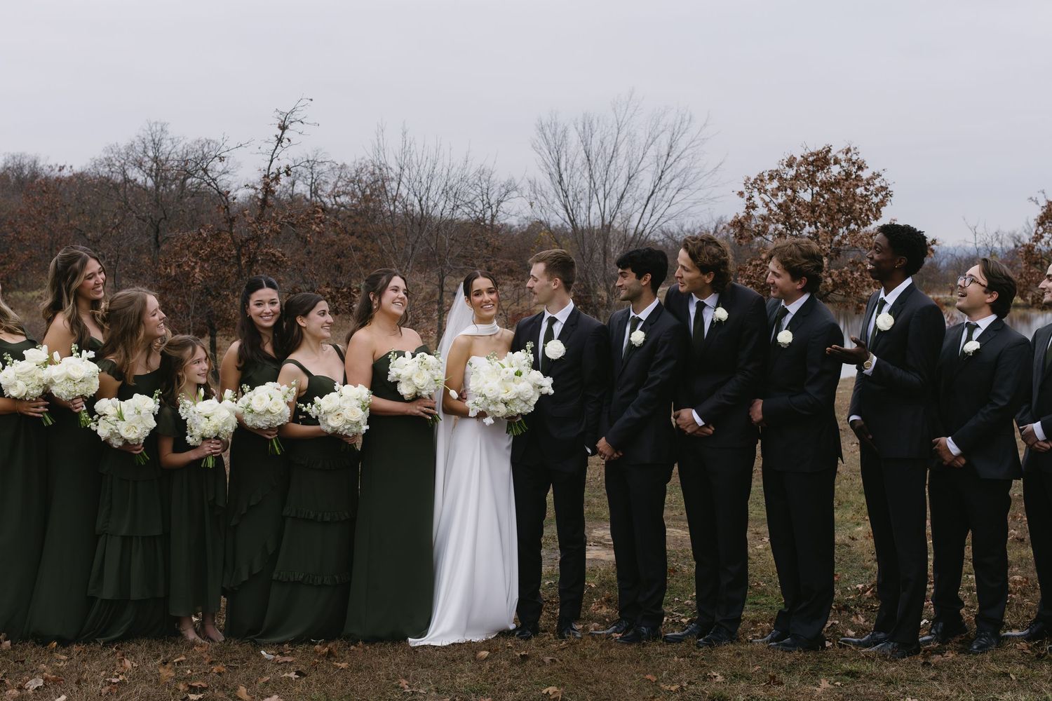 Wedding party lineup outdoors with bridesmaids in black dresses against an autumn landscape.