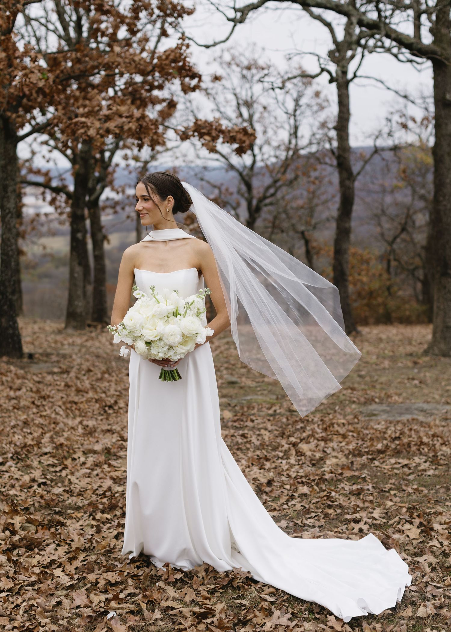 An elegant bride in a strapless white gown holds white bouquet among autumn trees.