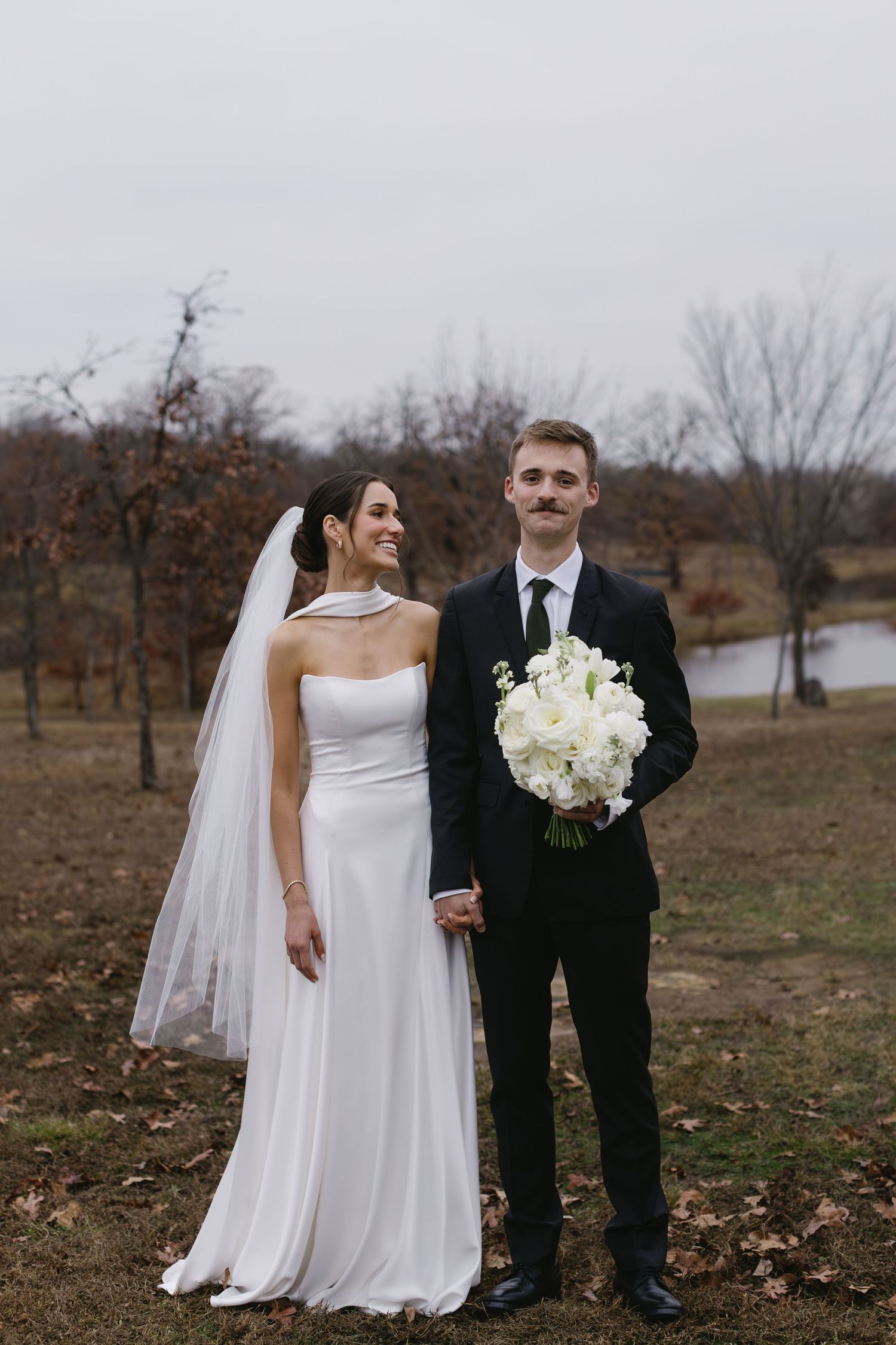 Wedding party poses in formal attire on autumn grounds with bare trees and fallen leaves.
