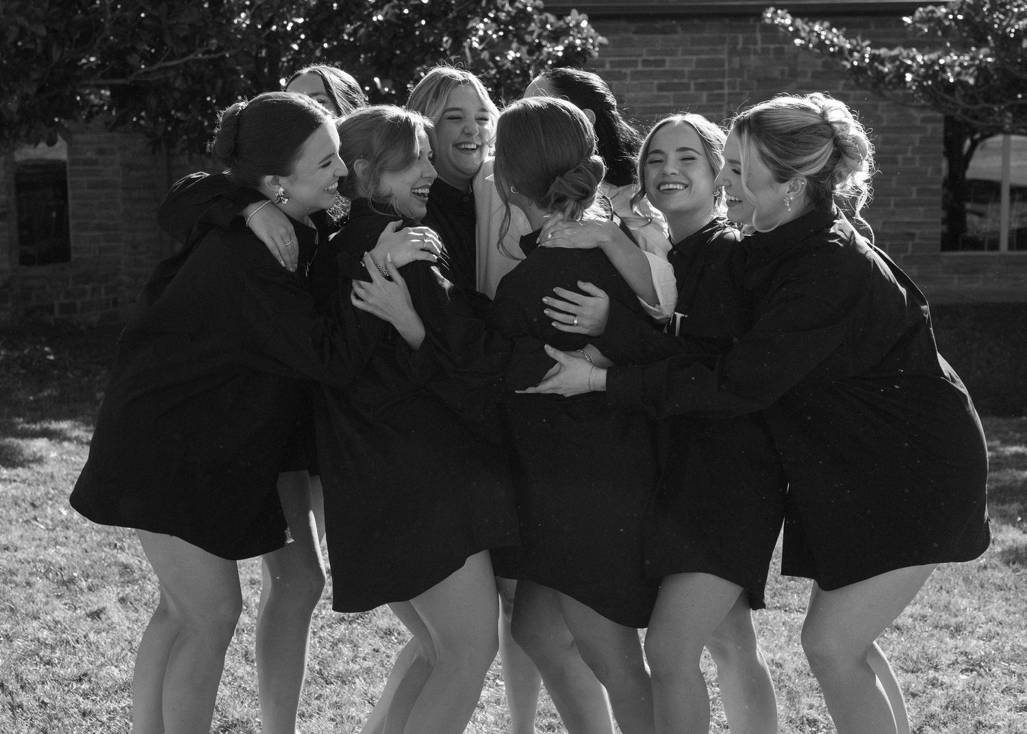Wedding party in black dresses embracing outdoors in a candid moment.