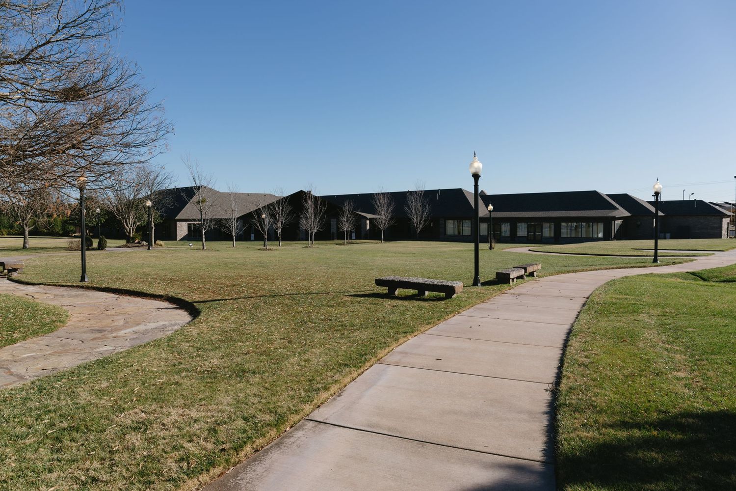 Concrete pathway winding through a grassy park area with benches and buildings in the background.