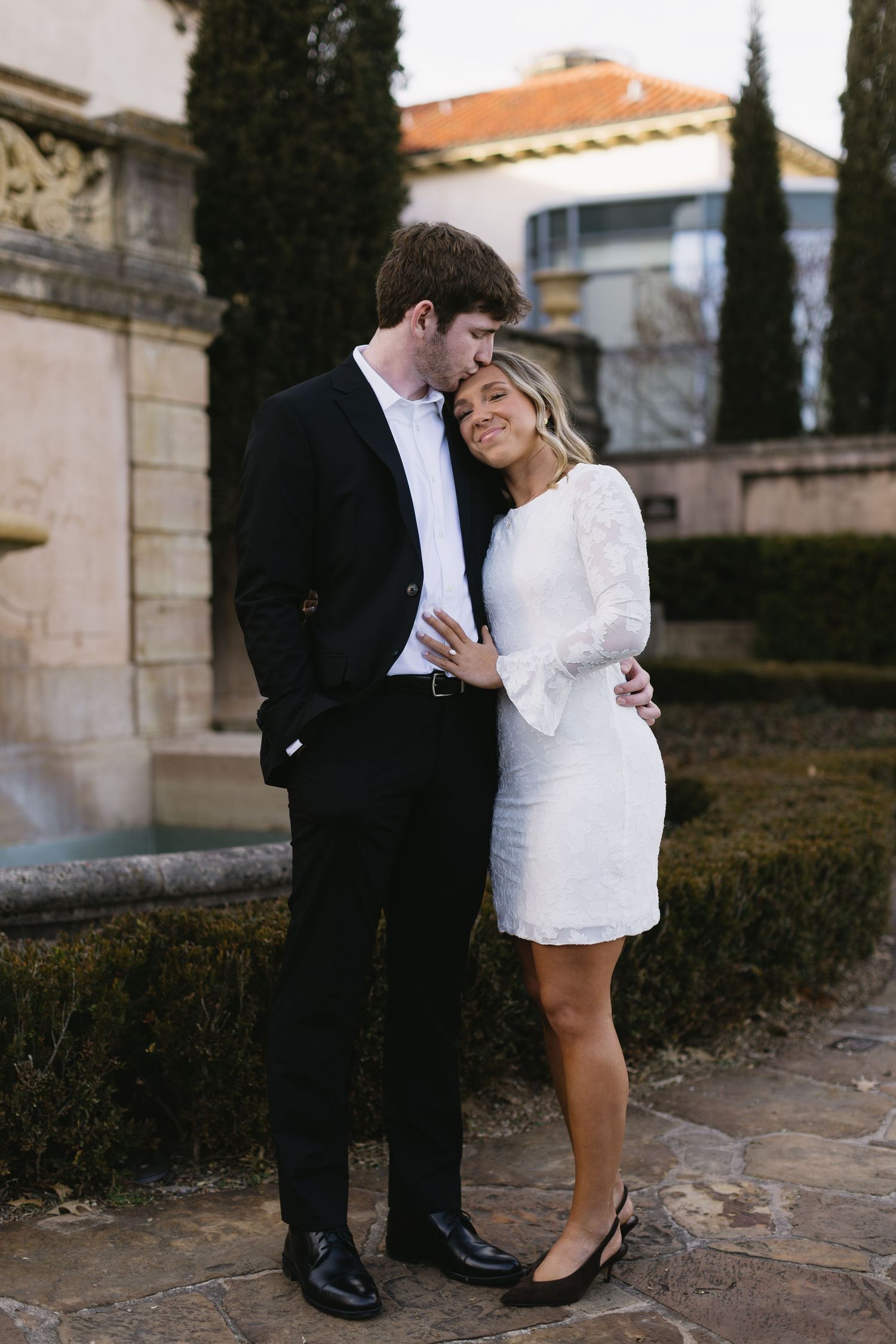 A couple embraces in formal attire near a Mediterranean-style building with cypress trees.