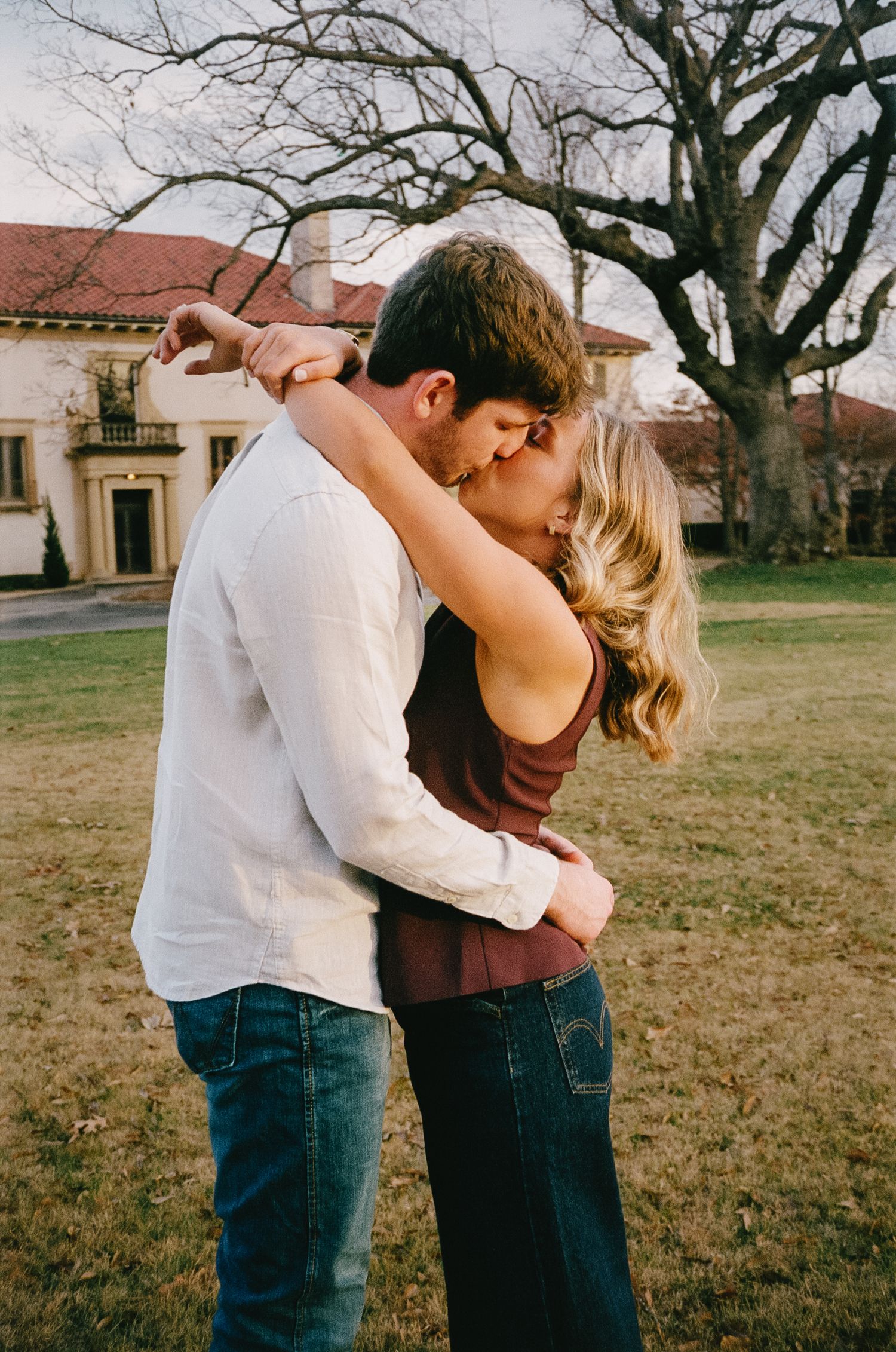 A couple shares a passionate kiss on a lawn with Mediterranean-style architecture in the background.