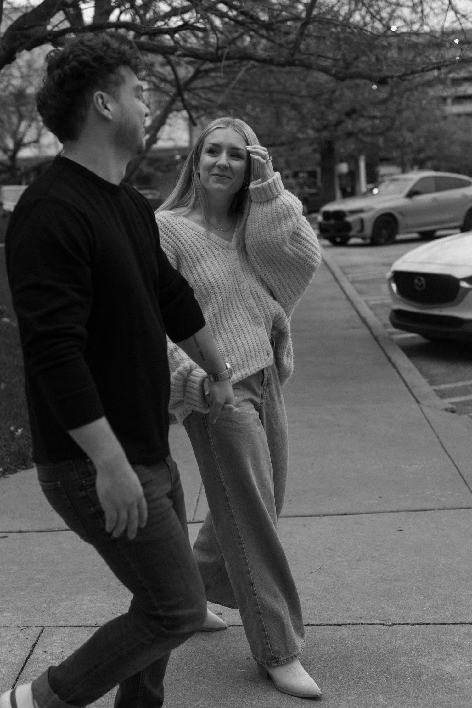 A couple shares a playful moment on a sidewalk in black and white photography.