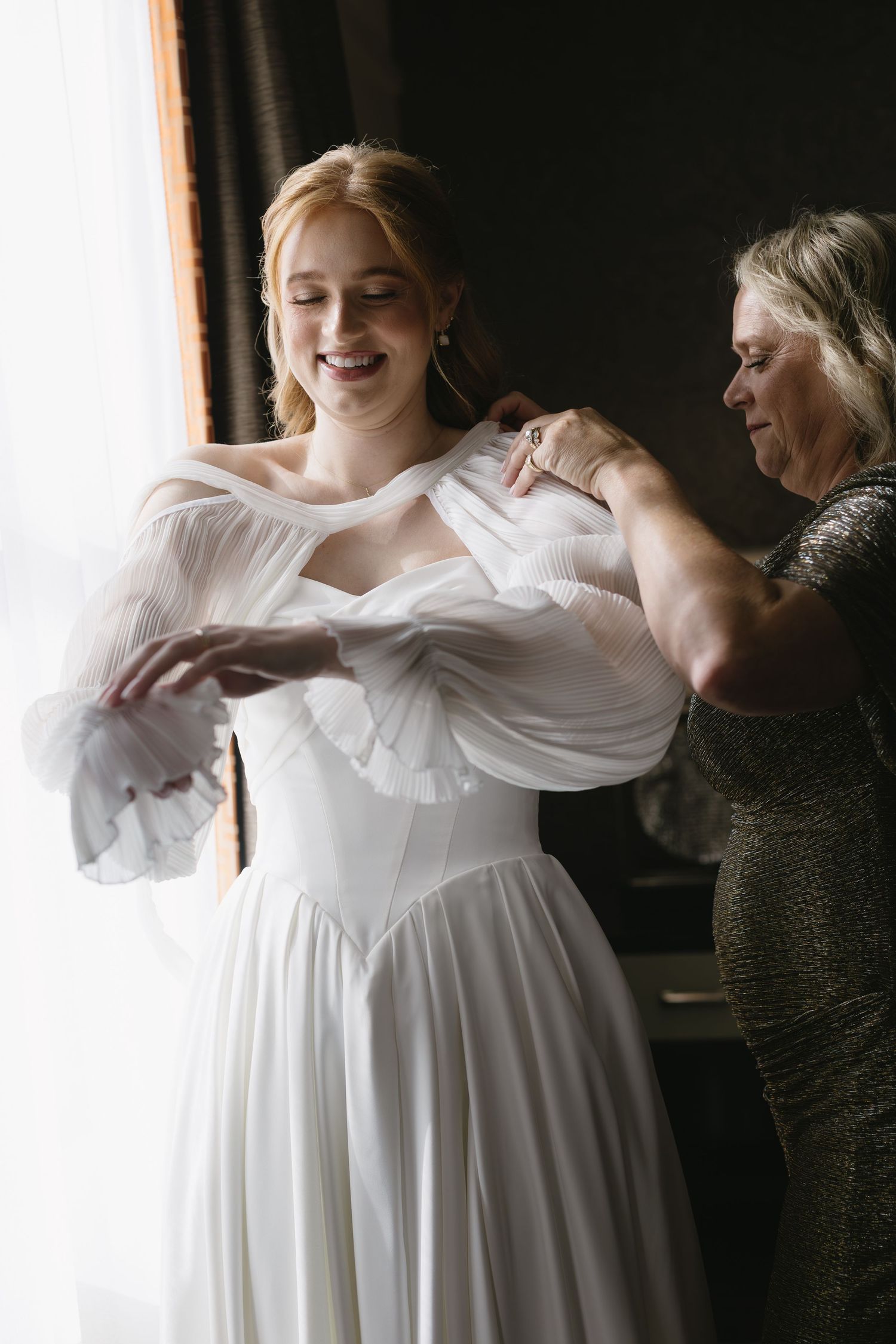 A tender moment during wedding preparations as a dress is adjusted near a bright window.