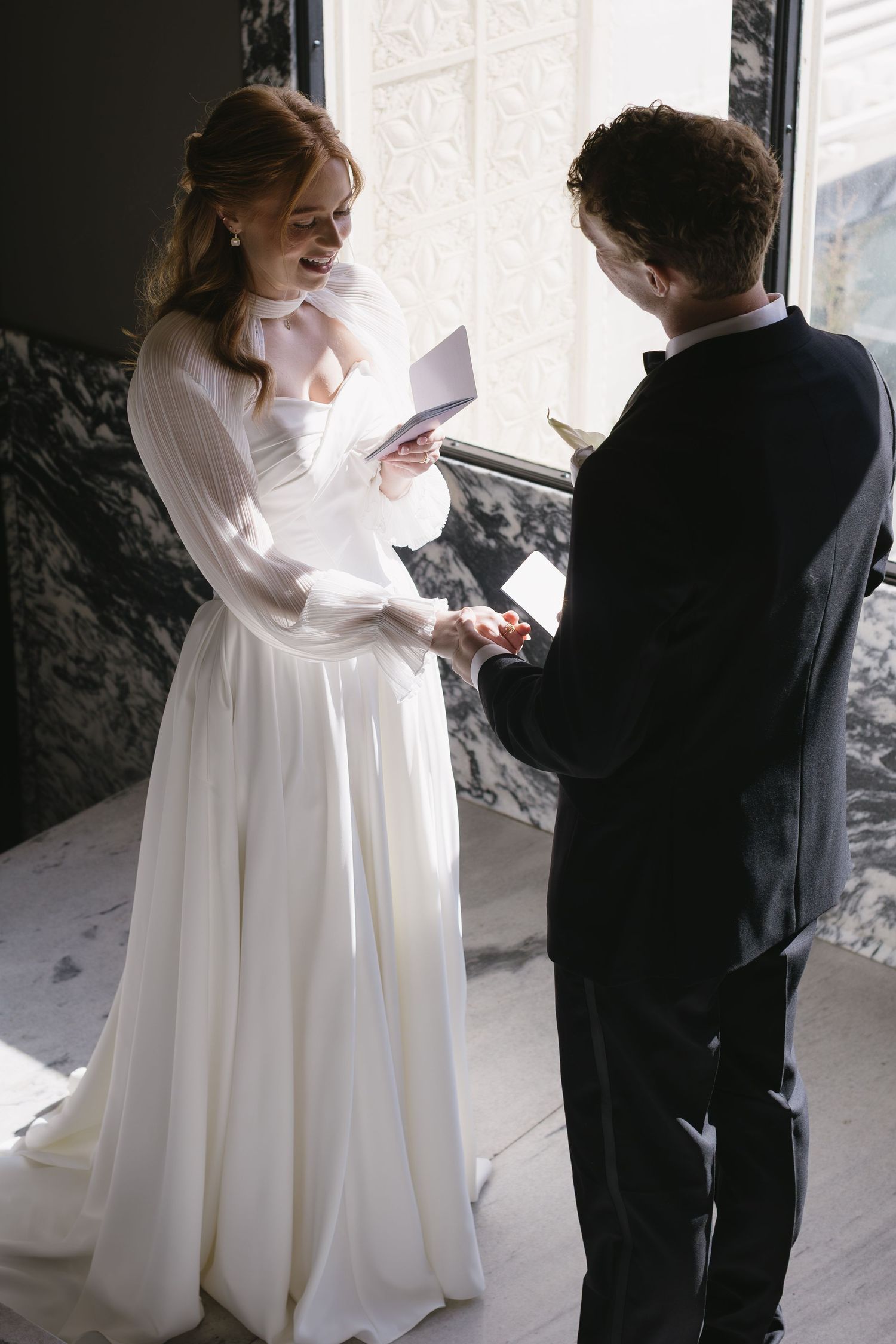 Wedding couple exchanging items in a sunlit corridor with dramatic lighting.