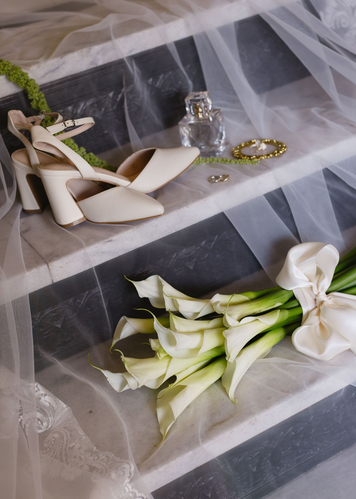 Wedding accessories including white heels and calla lily flowers arranged on white tulle fabric.