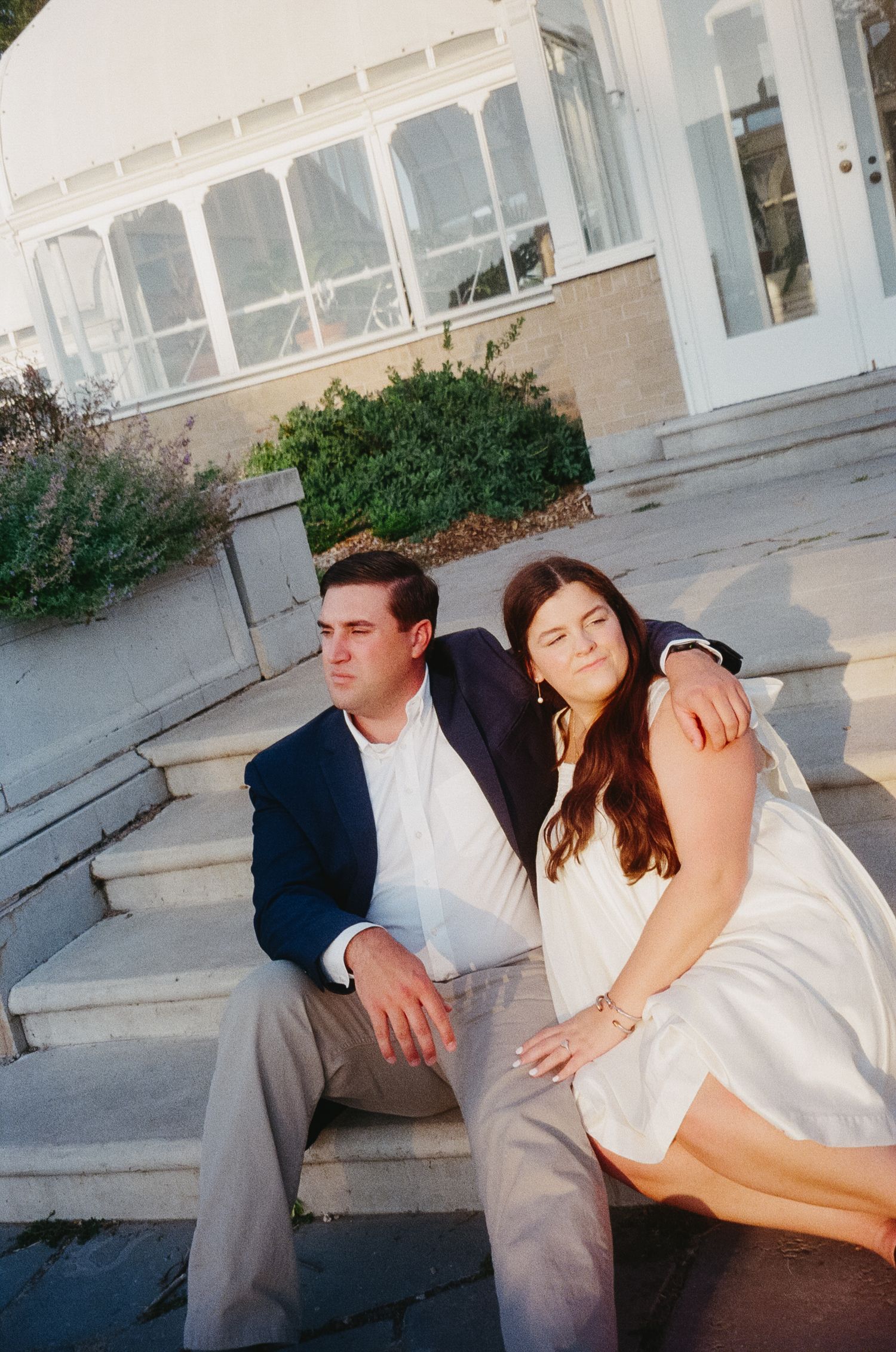 A couple sits together on concrete steps outside a white building with windows.