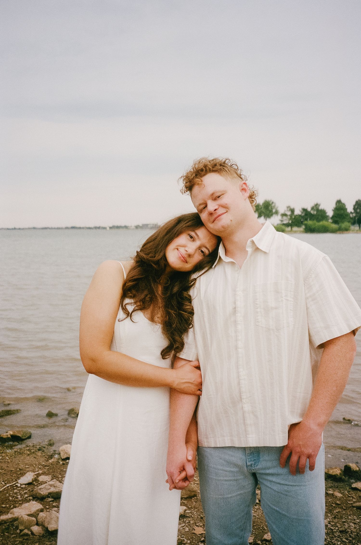 A couple in casual white attire poses lovingly together by a waterfront on a cloudy day.