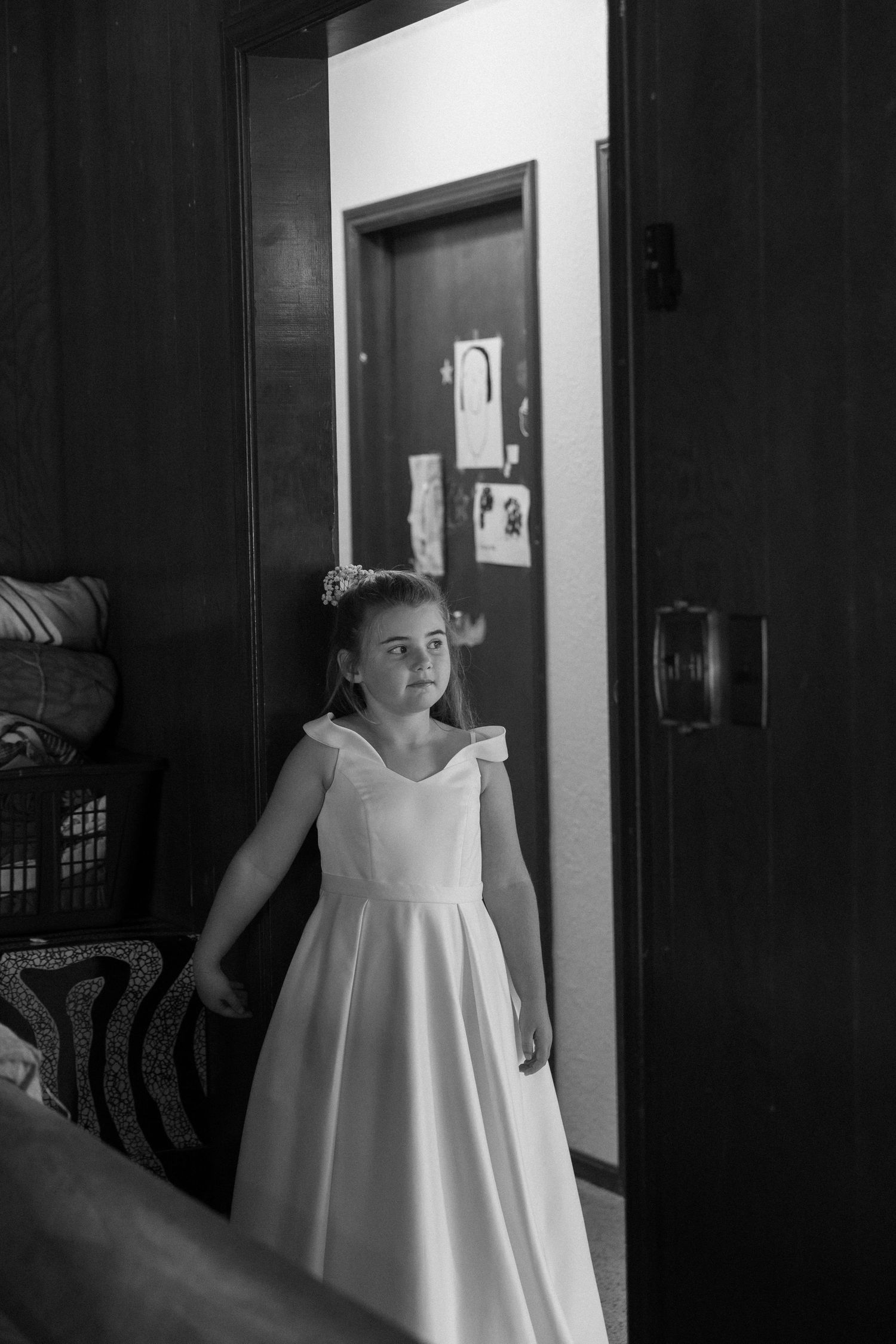 Young flower girl in a white sleeveless dress standing in a dimly lit hallway.