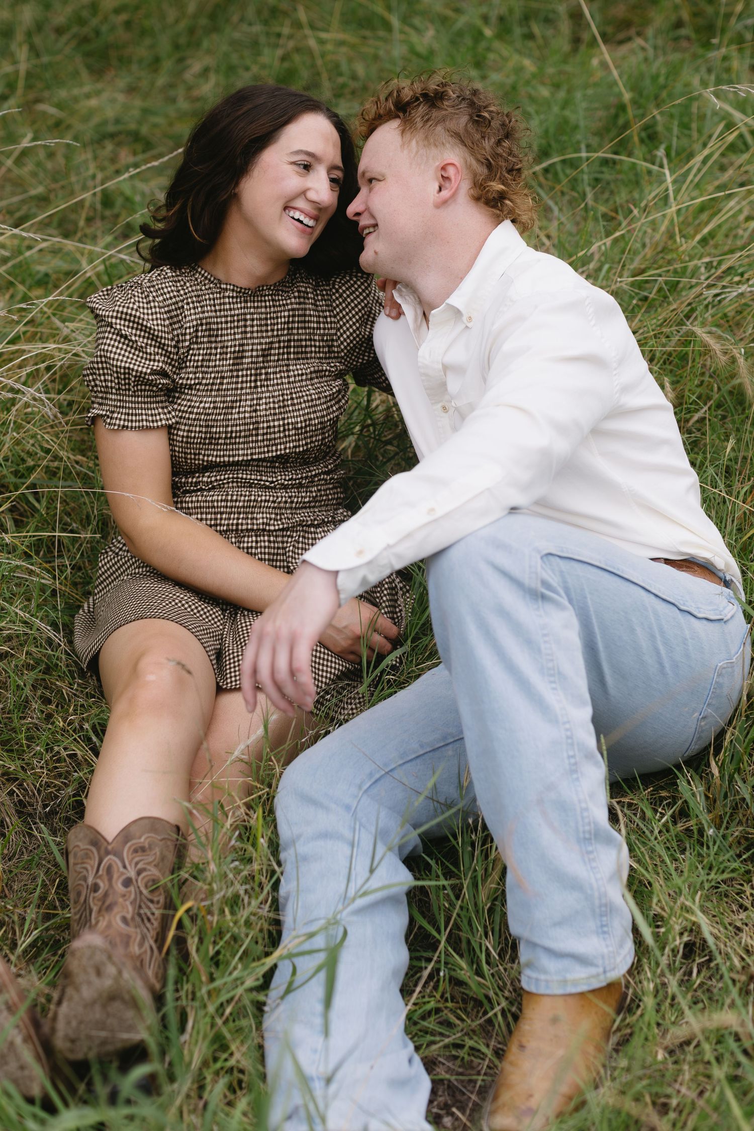 Loving couple lounges together in tall grass wearing cowboy boots and casual attire.