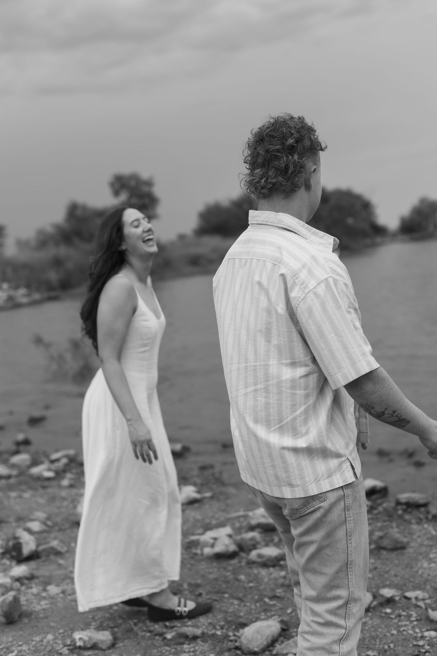 A couple in white and beige outfits poses playfully on a rocky beach in black and white photography.