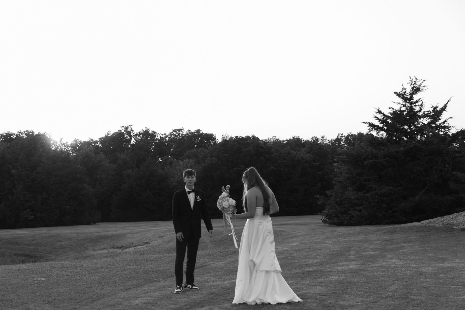 Elegant wedding couple walks across sprawling green lawn at sunset with trees in background.