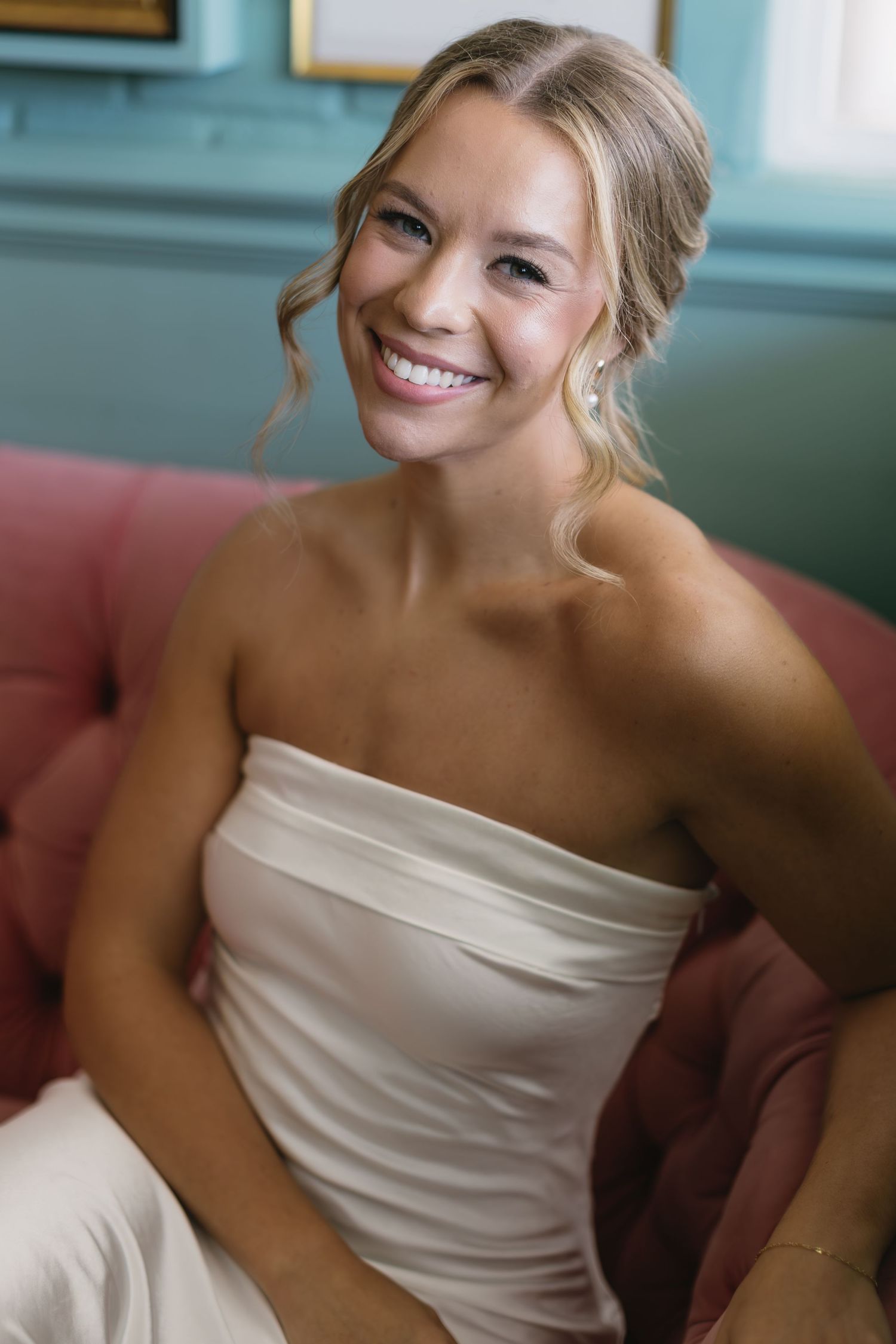 Joyful bride wearing strapless dress lounges on pink sofa during wedding day preparations.