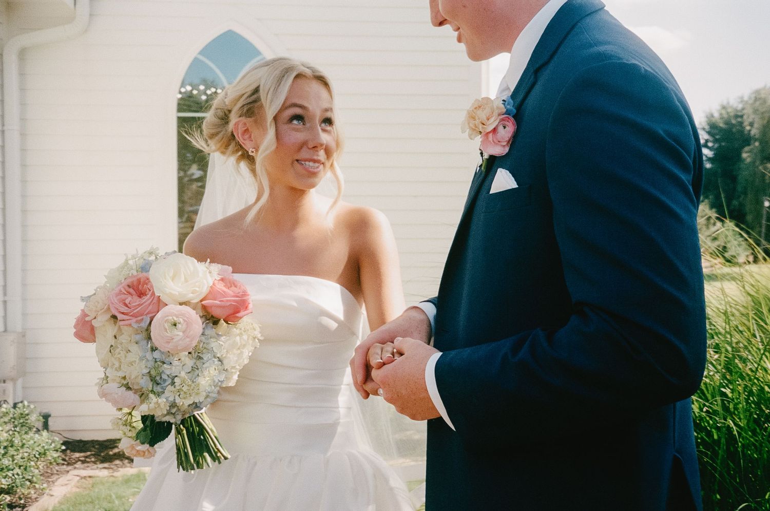 A couple on their wedding day standing close together with a blush and white rose bouquet against a sunny outdoor backdrop.