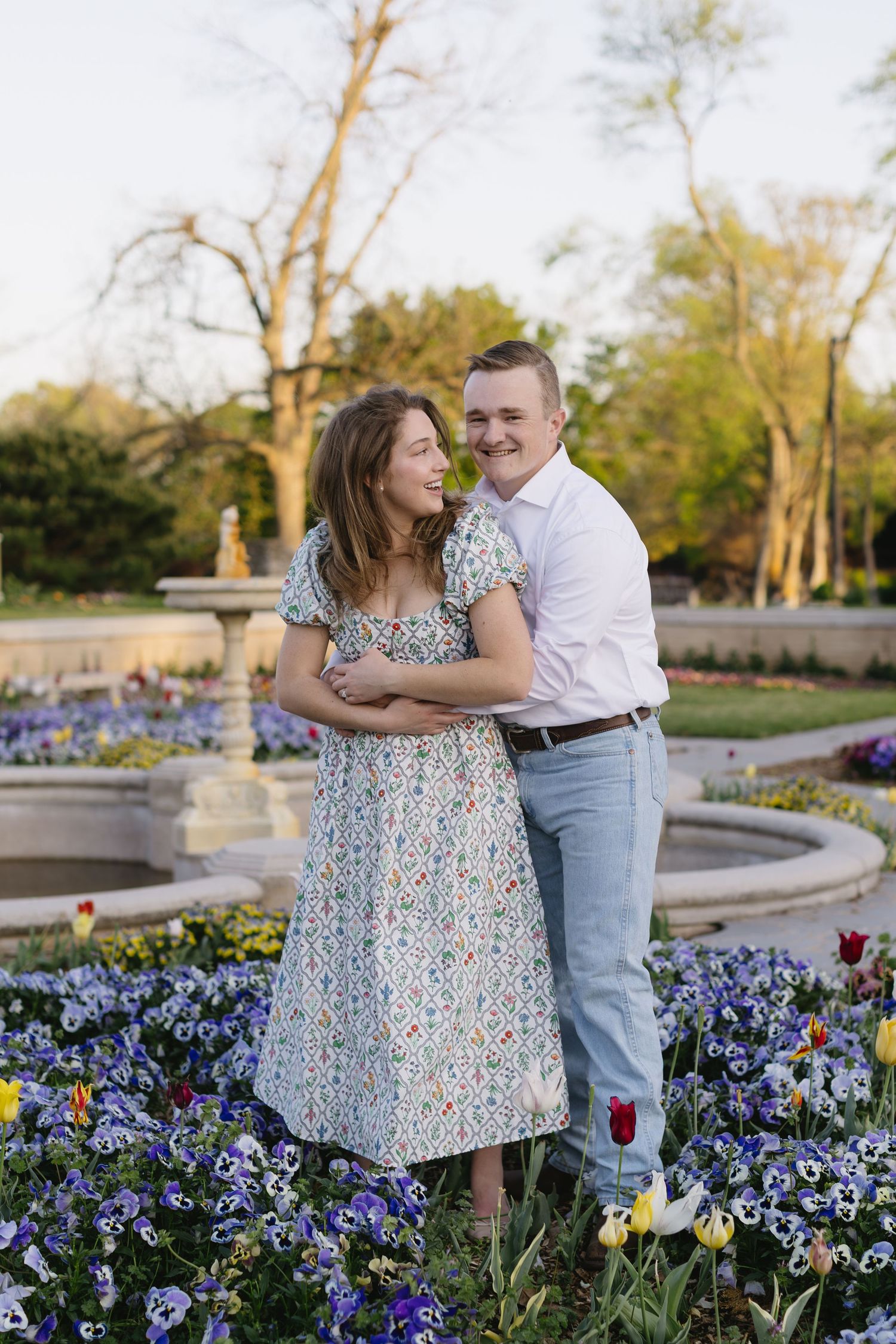 A loving couple poses together in a garden filled with colorful tulips and purple flowers at sunset.