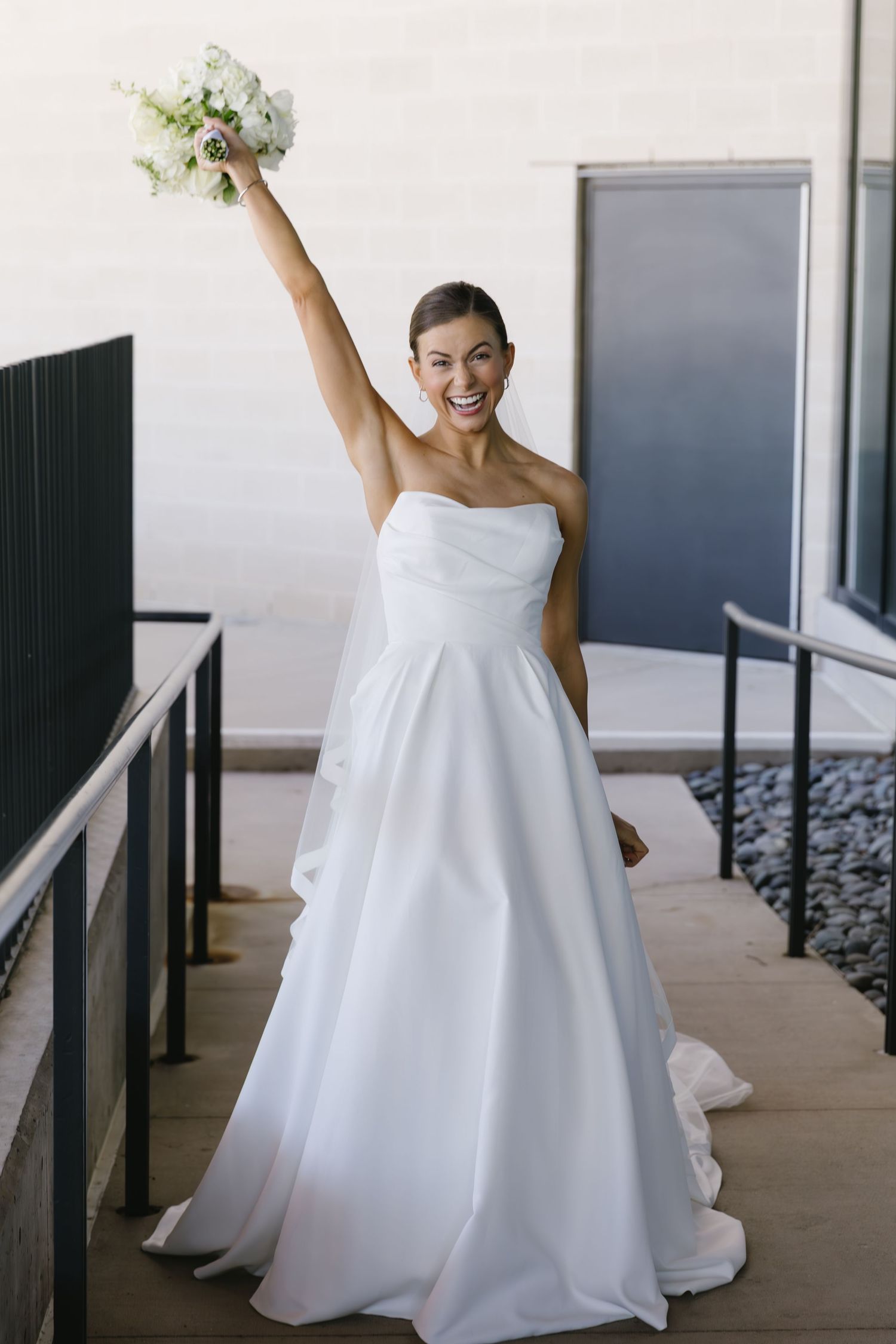 A bride in a strapless white wedding gown raises her bouquet joyfully on an outdoor terrace.