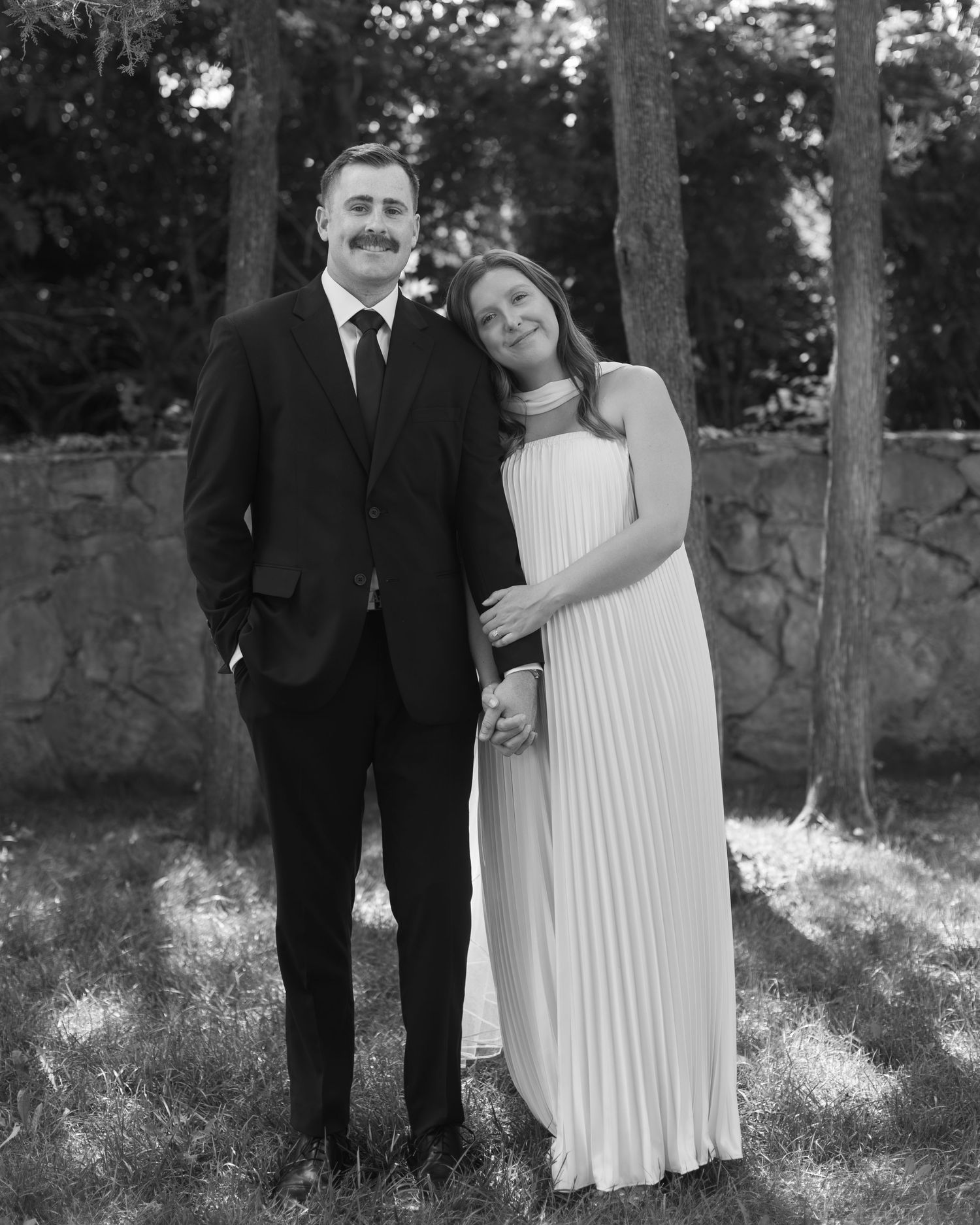 Black and white wedding photos showing a couple in formal attire posing together outdoors by stone walls and trees.