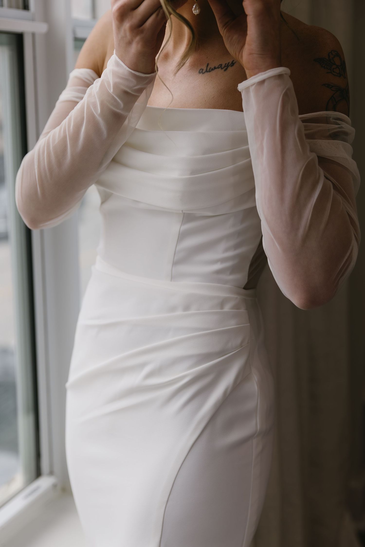 Close-up of white wedding dress with sheer sleeves near window.