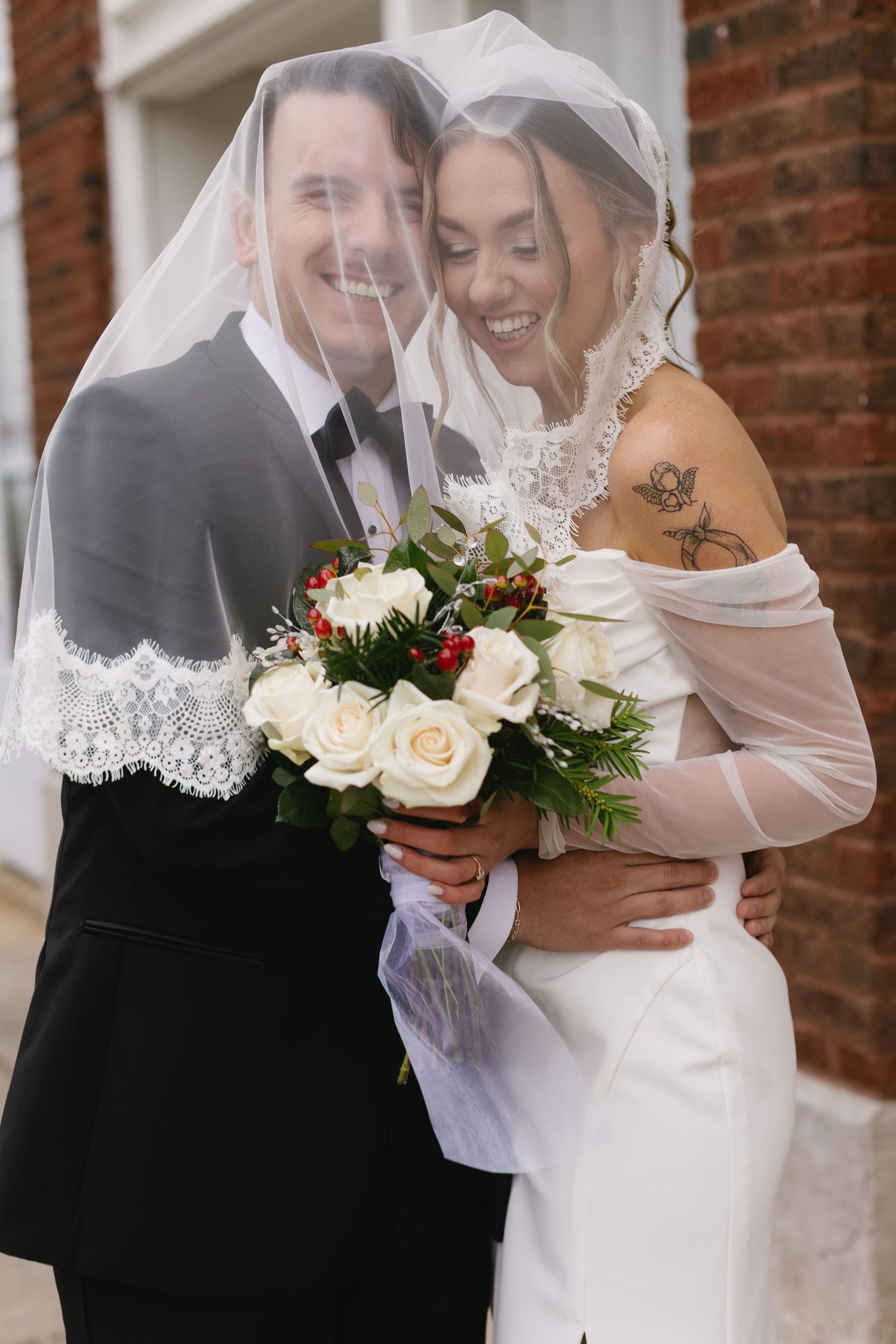 Joyful wedding couple sharing a laugh under a veil with white rose bouquet.
