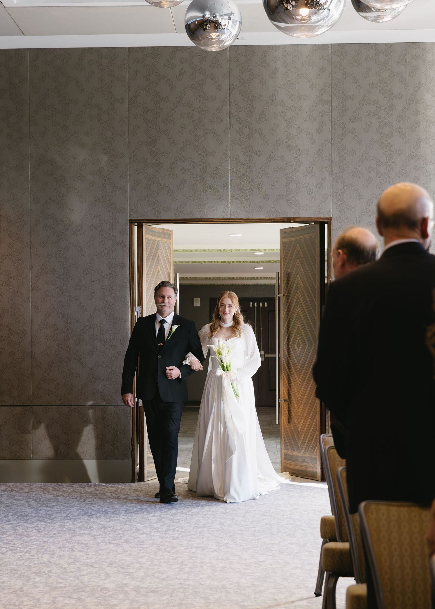 Wedding party walking through an elegant ballroom with hanging disco balls and decorative lighting.