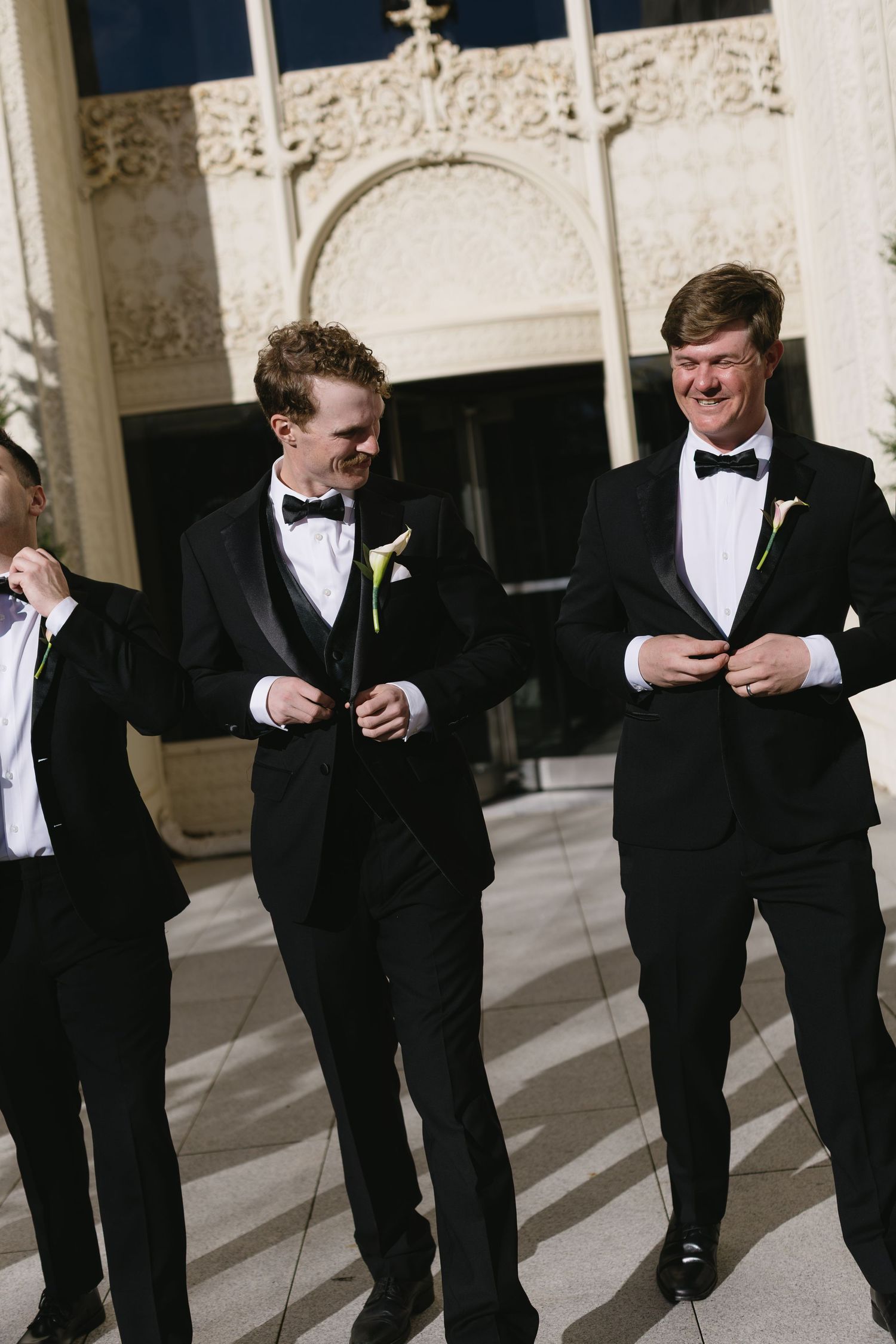 Groomsmen in classic black tuxedos share a candid moment outside the wedding venue.