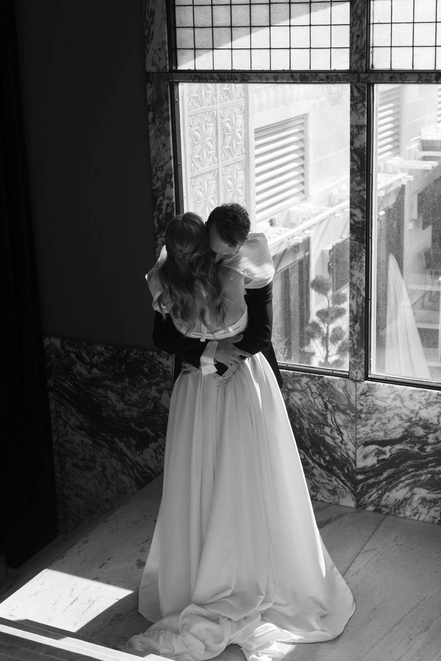 A romantic black and white wedding photo of a couple embracing by a window in formal attire.