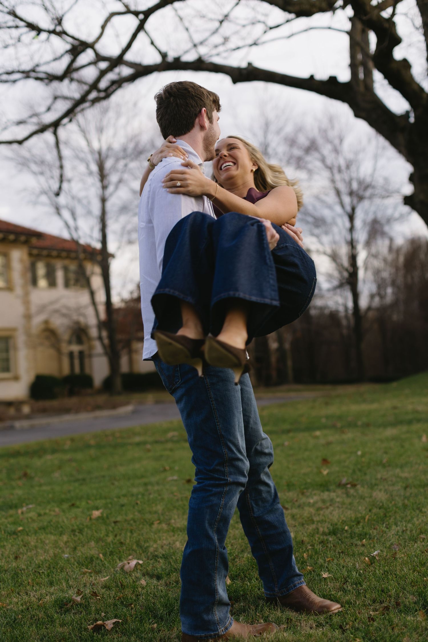 A romantic couple shares intimate moments on a grassy lawn with bare trees and buildings in the background.