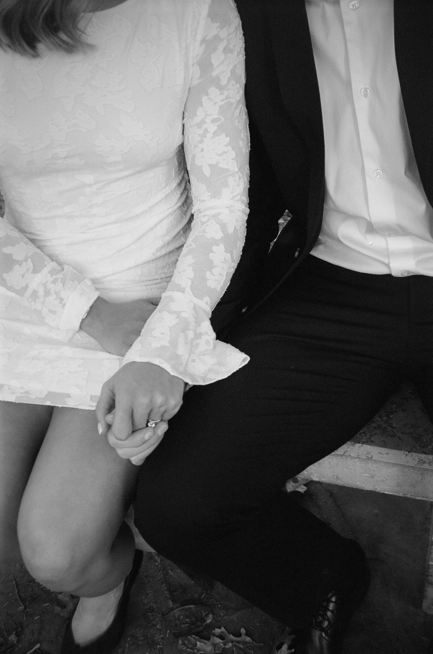 A close-up detail shot of hands and formal attire, featuring a lace wedding dress and dark suit.