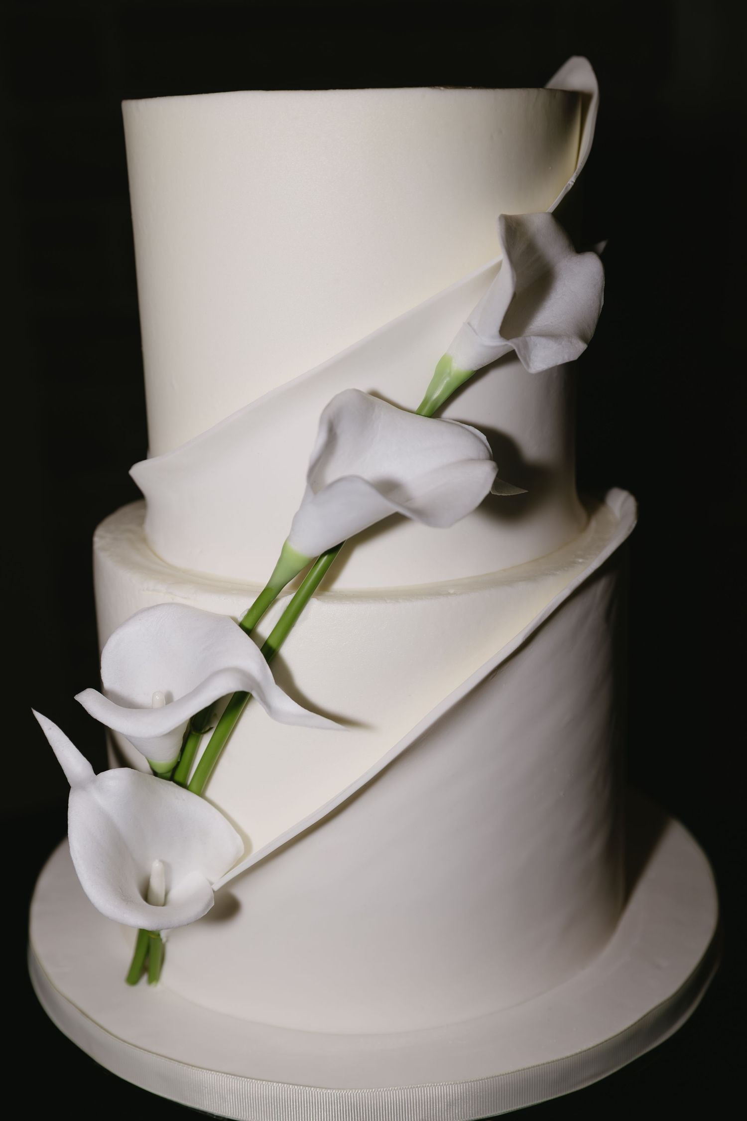 Elegant white wedding cake decorated with delicate calla lilies against black background.
