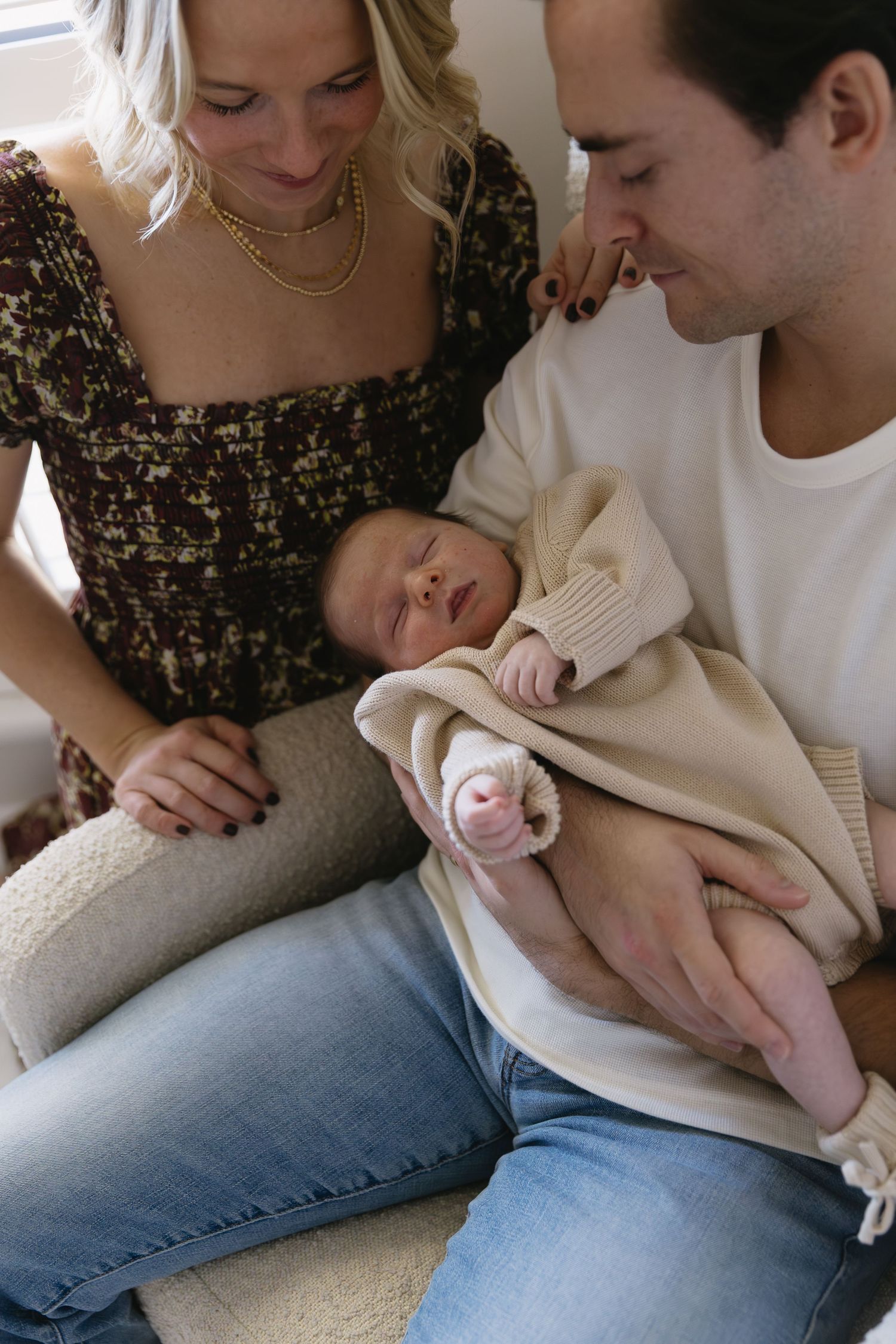 A couple tenderly holds their newborn baby wrapped in a beige knit blanket while sitting together on a couch.