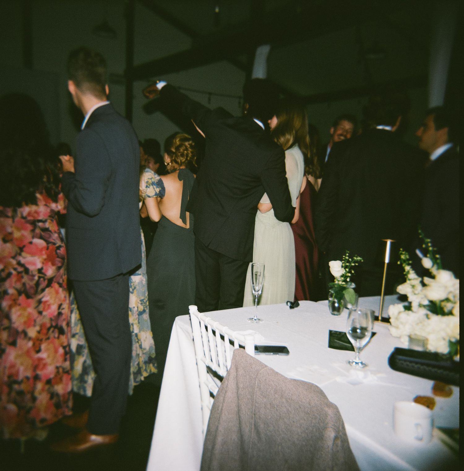 Wedding reception guests gathered around elegant white-clothed dining tables in a dimly lit venue.