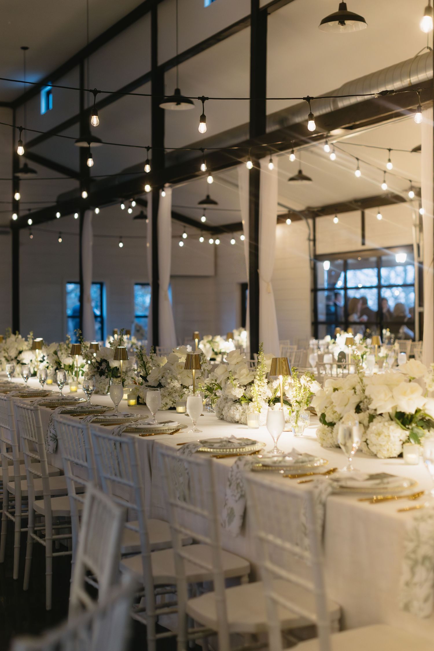 Modern reception venue with white tables and chairs decorated with string lights and floral centerpieces.