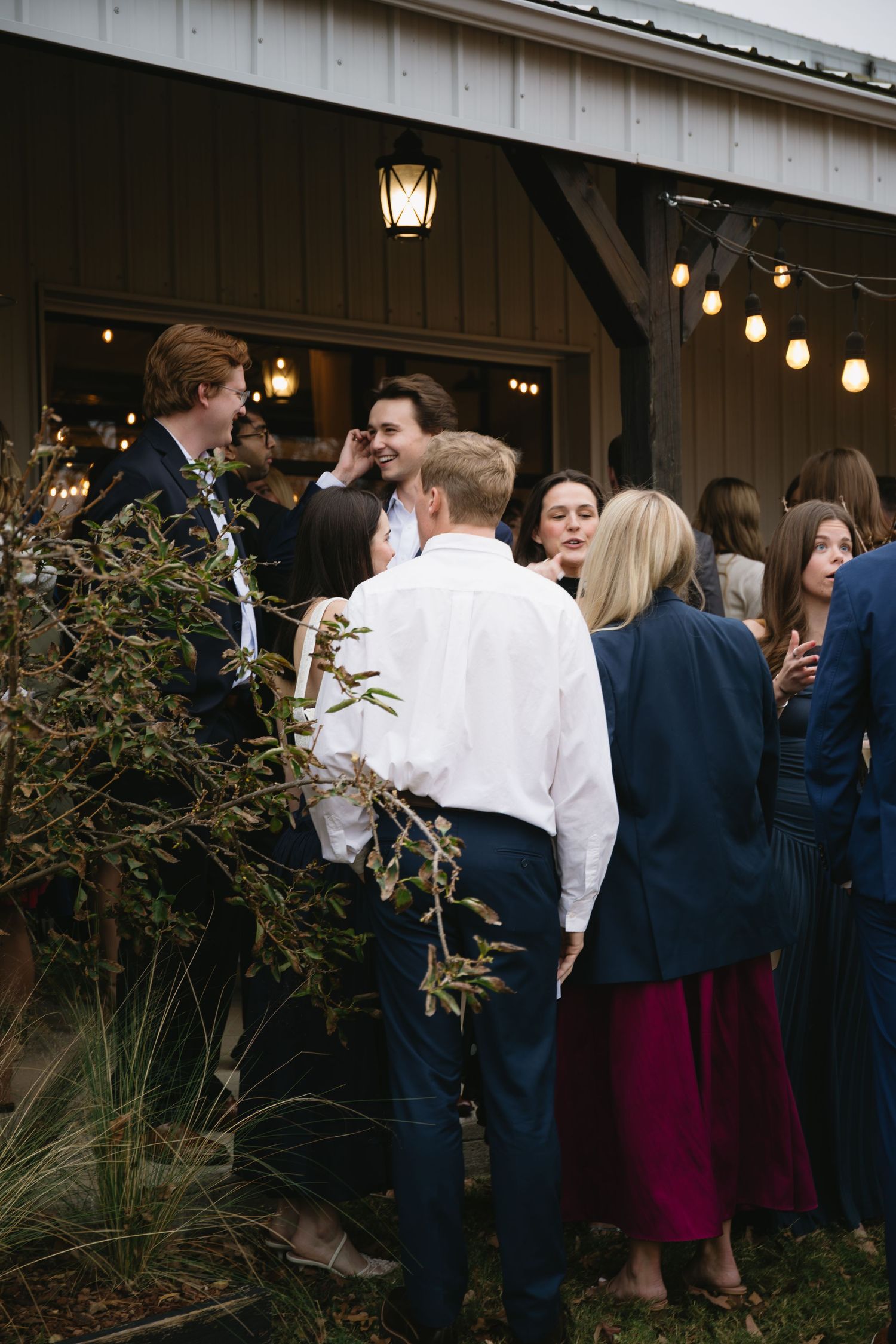 Wedding guests gather outside a white venue decorated with string lights at dusk.