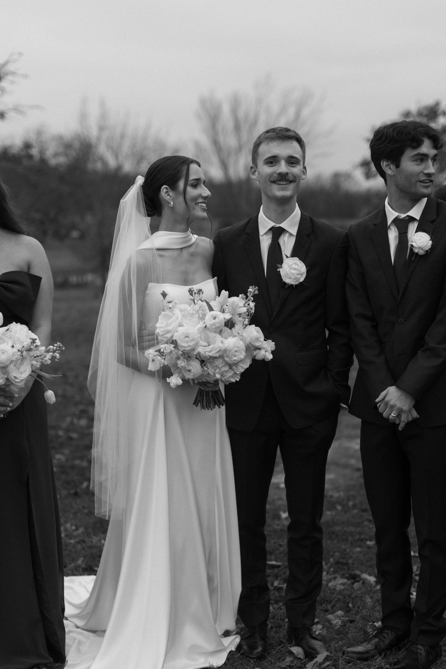 Wedding party poses outdoors in formal attire with the bride holding a white bouquet.
