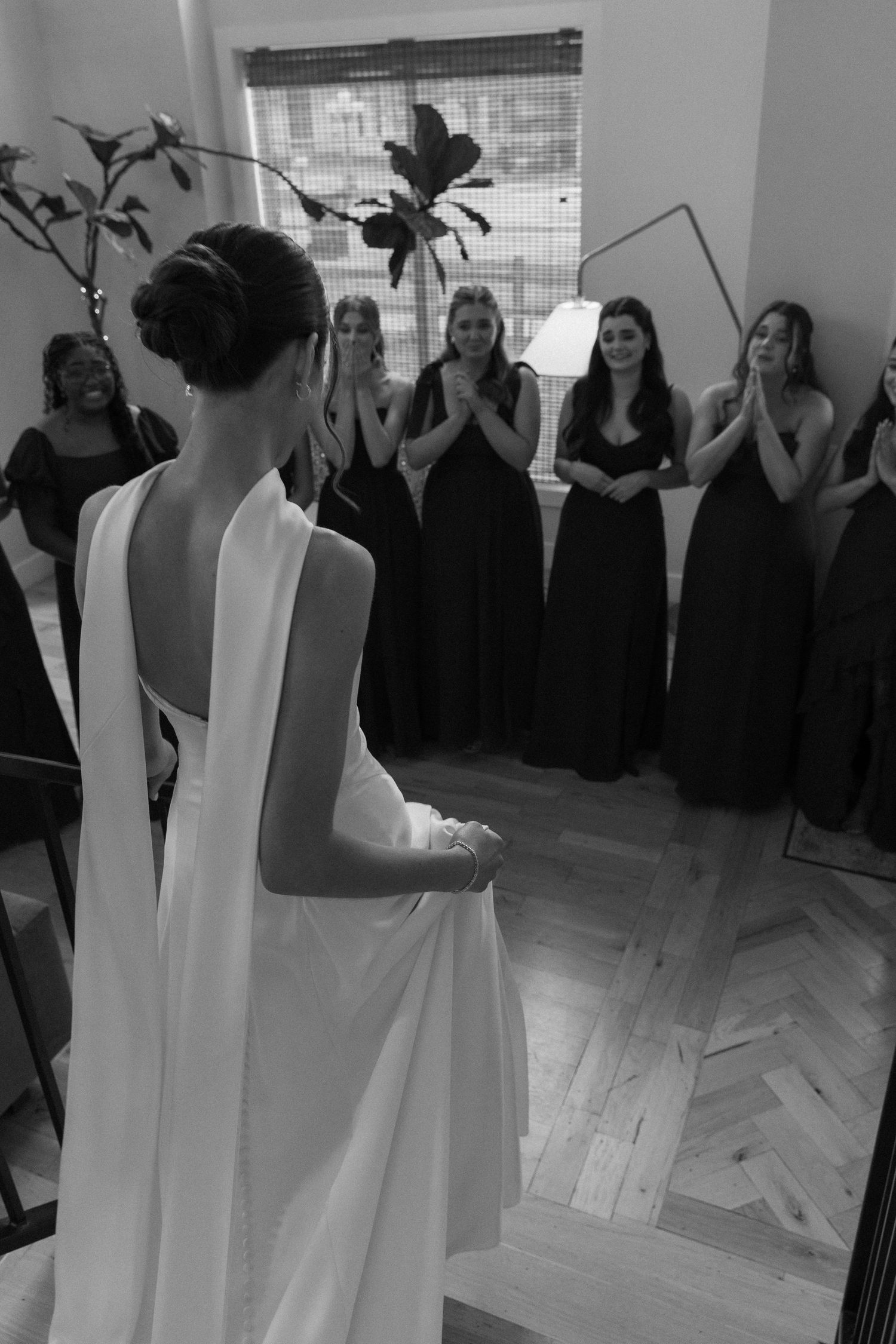 Bridesmaids in black dresses react with joy as they see the bride in her elegant white gown.