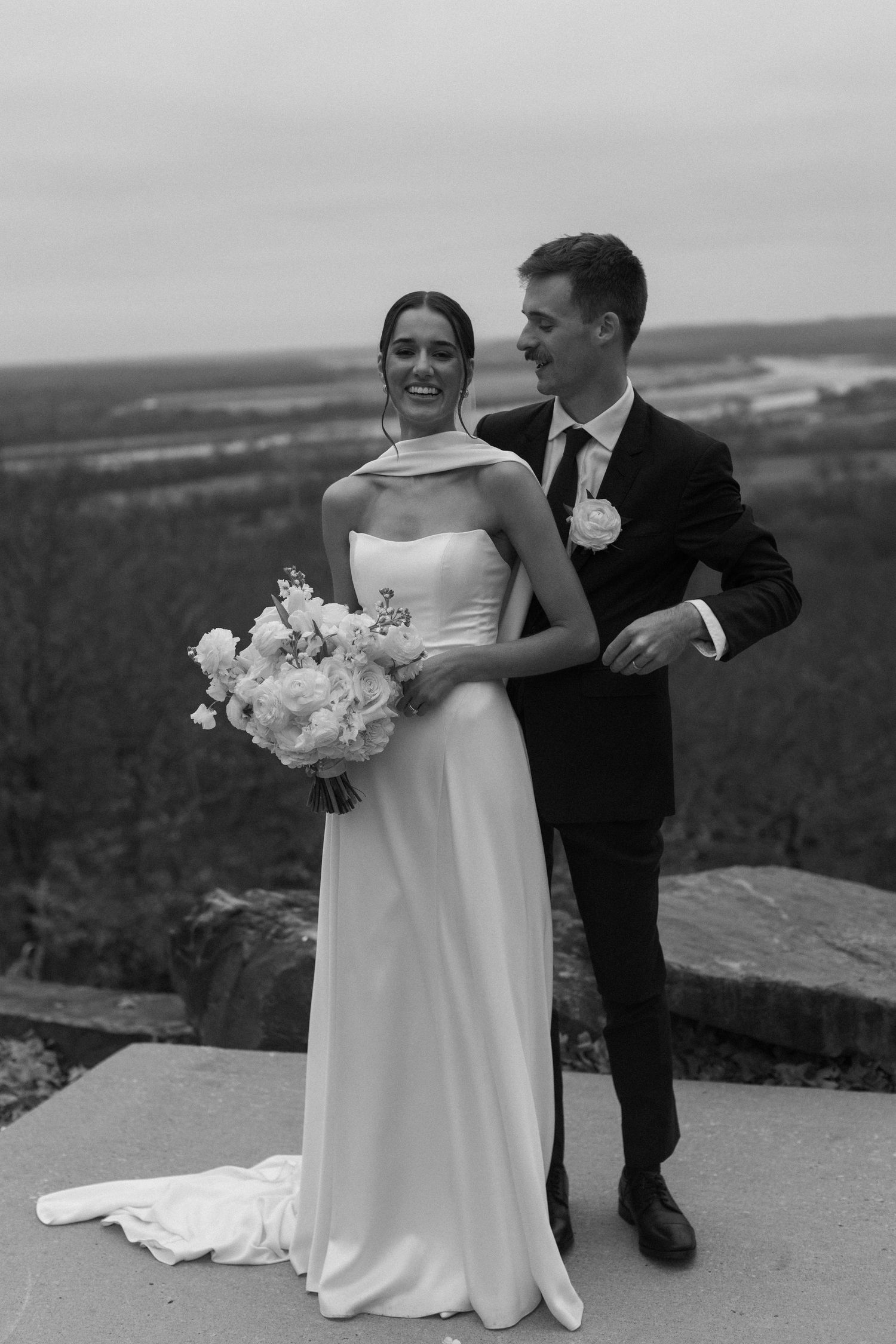 A couple in wedding attire share a romantic moment on a scenic overlook with white floral bouquet.