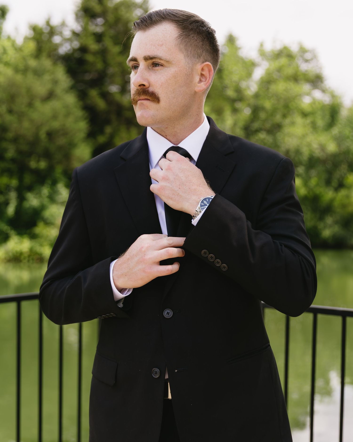 Groom adjusts his tie while wearing a classic black suit outdoors.