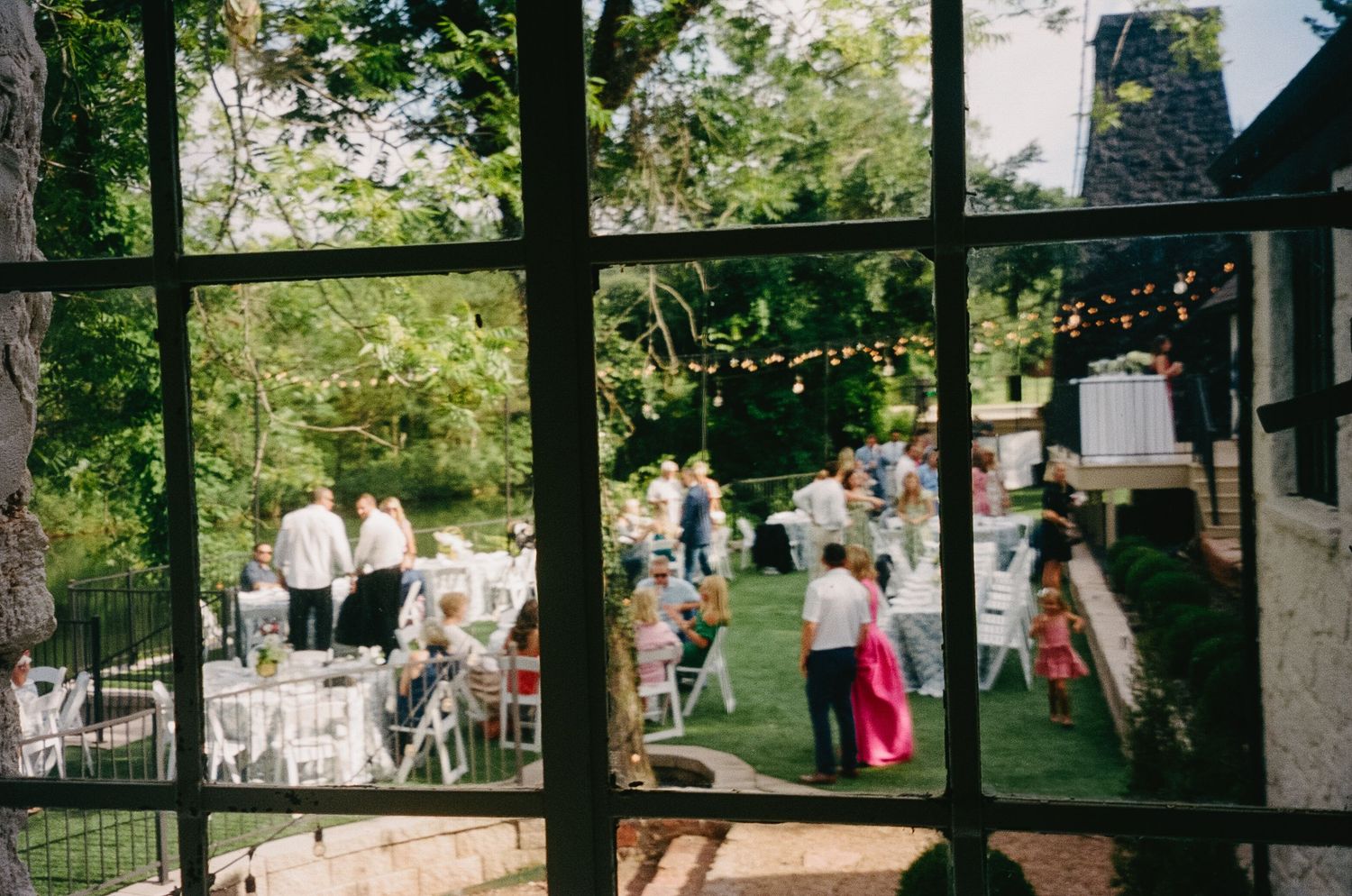 Wedding reception viewed through window panes with guests gathered under trees.