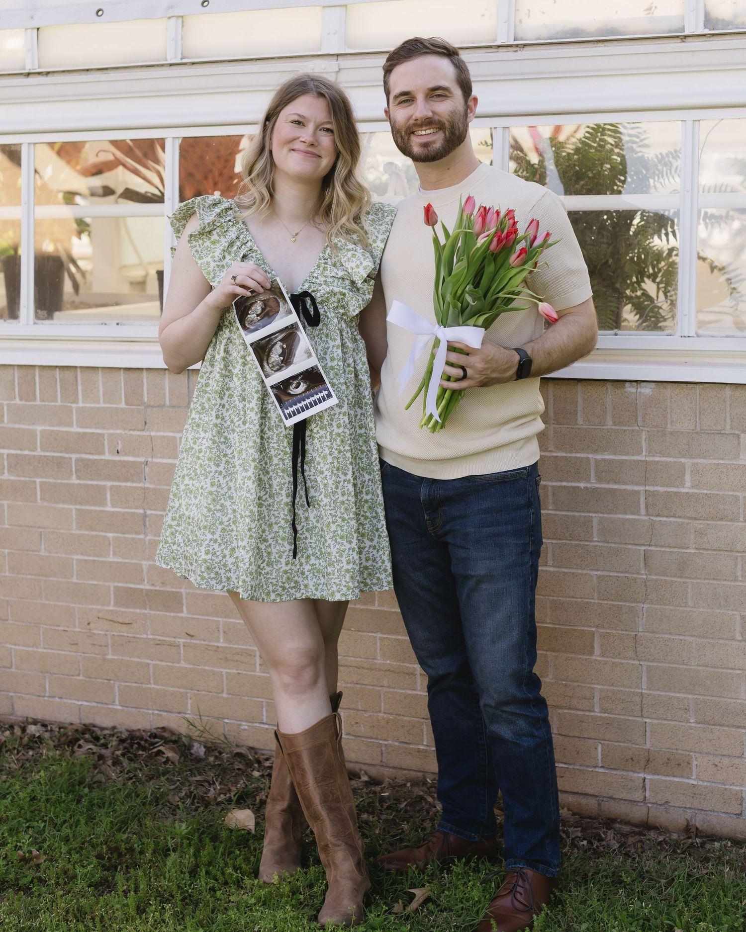 A couple celebrates a pregnancy announcement in front of a brick wall while holding red tulips and a sonogram photo.