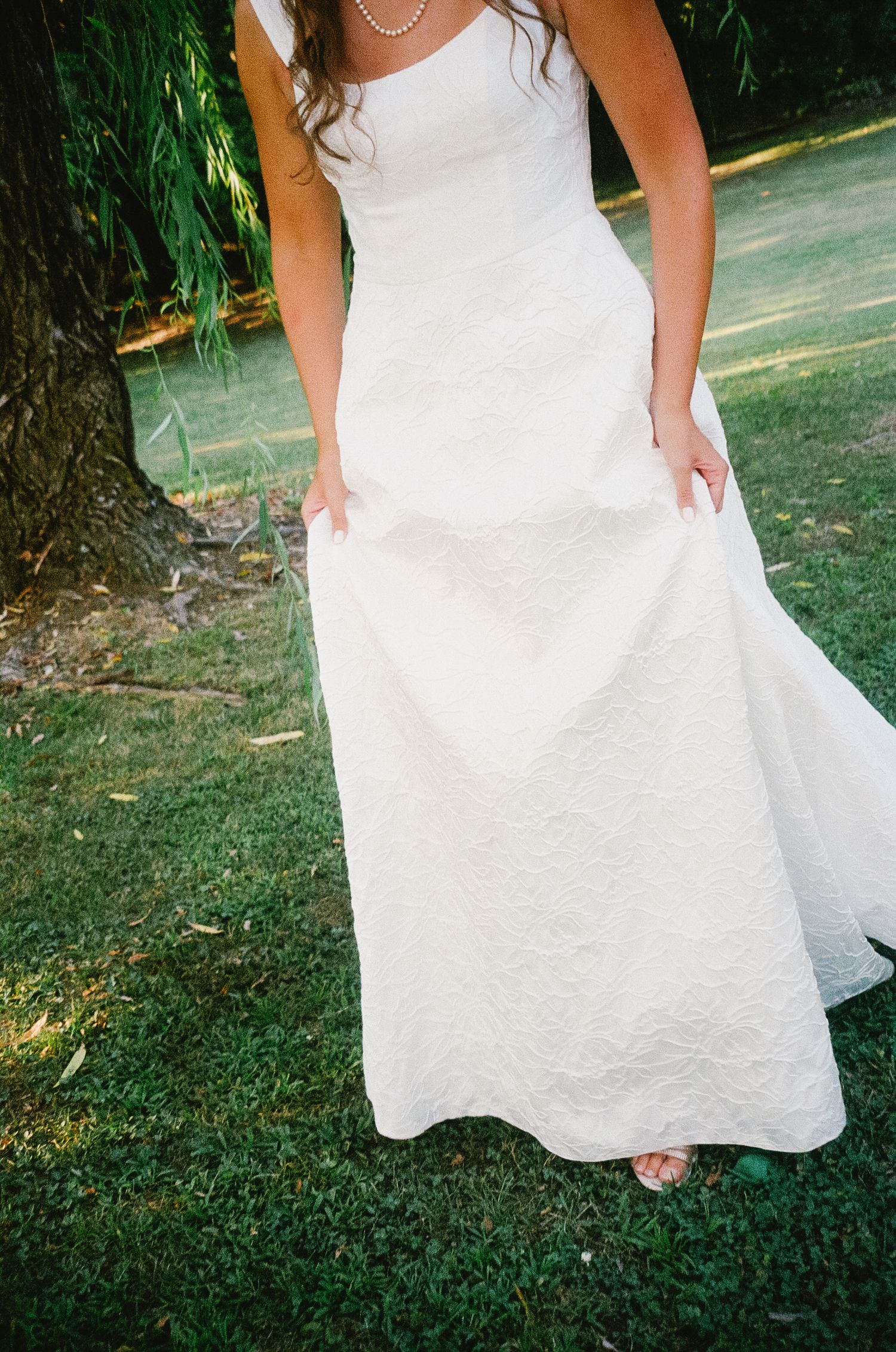 Detail shot of a white wedding dress skirt flowing against green grass in an outdoor garden setting.