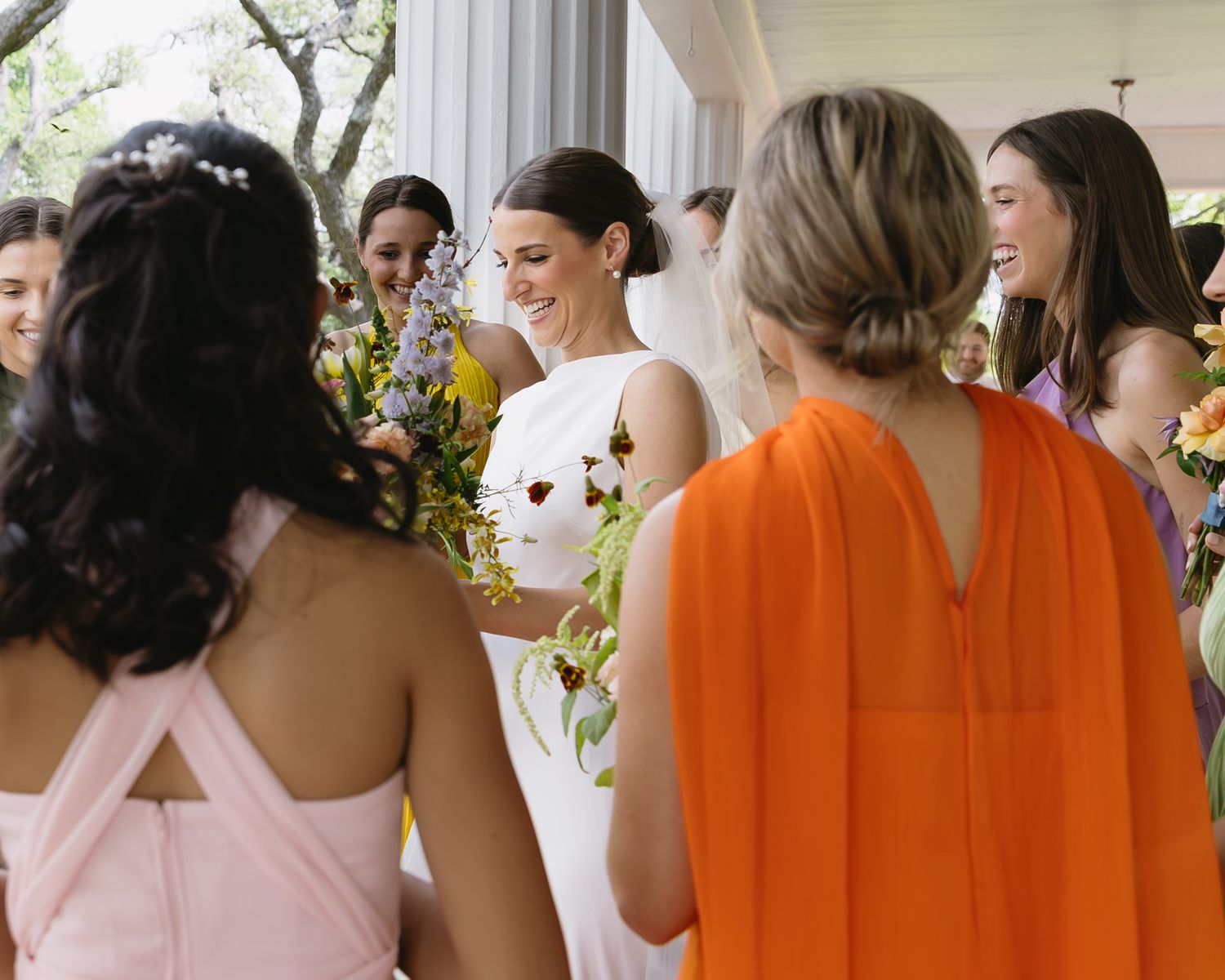 Row of bridesmaids in orange and pink dresses at outdoor wedding venue.