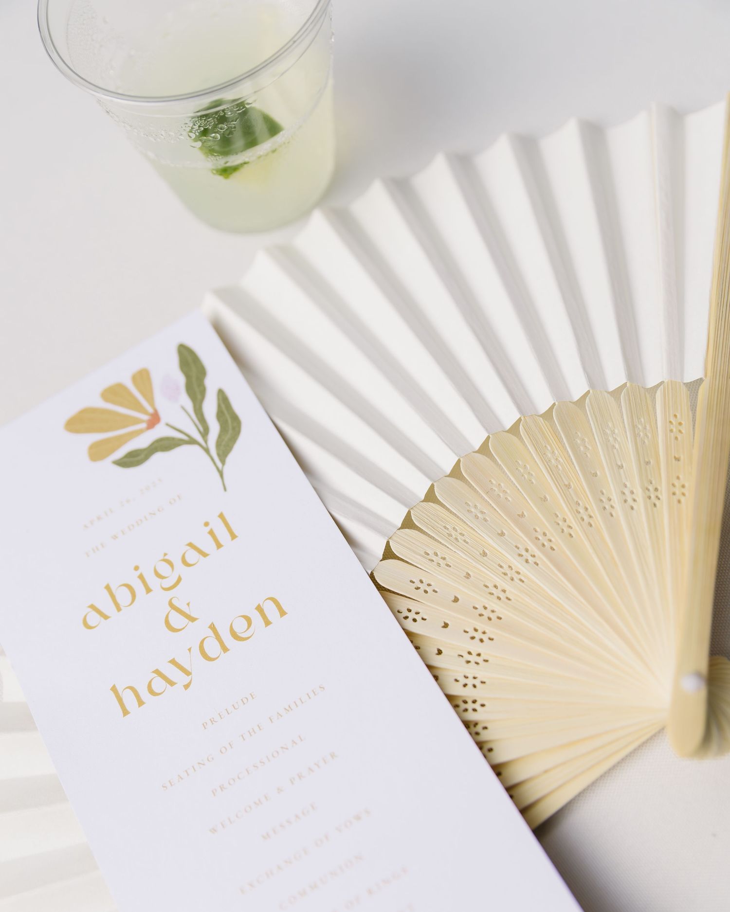 White paper fan and minimalist wedding invitation displayed with cocktail glasses and soft natural lighting.