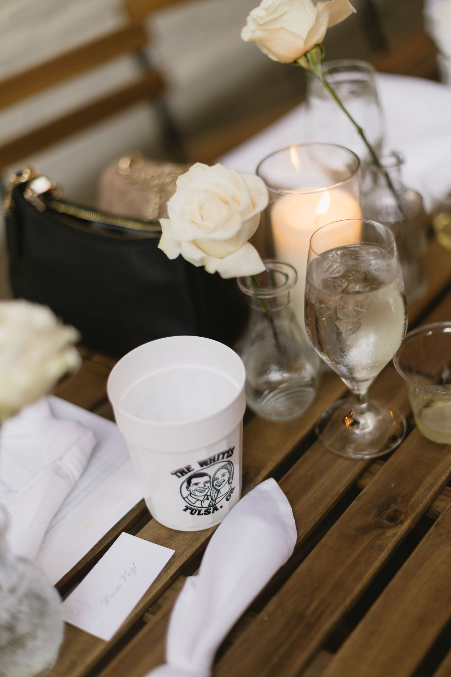 White roses and glassware arranged on a rustic wooden table setting with elegant place settings.