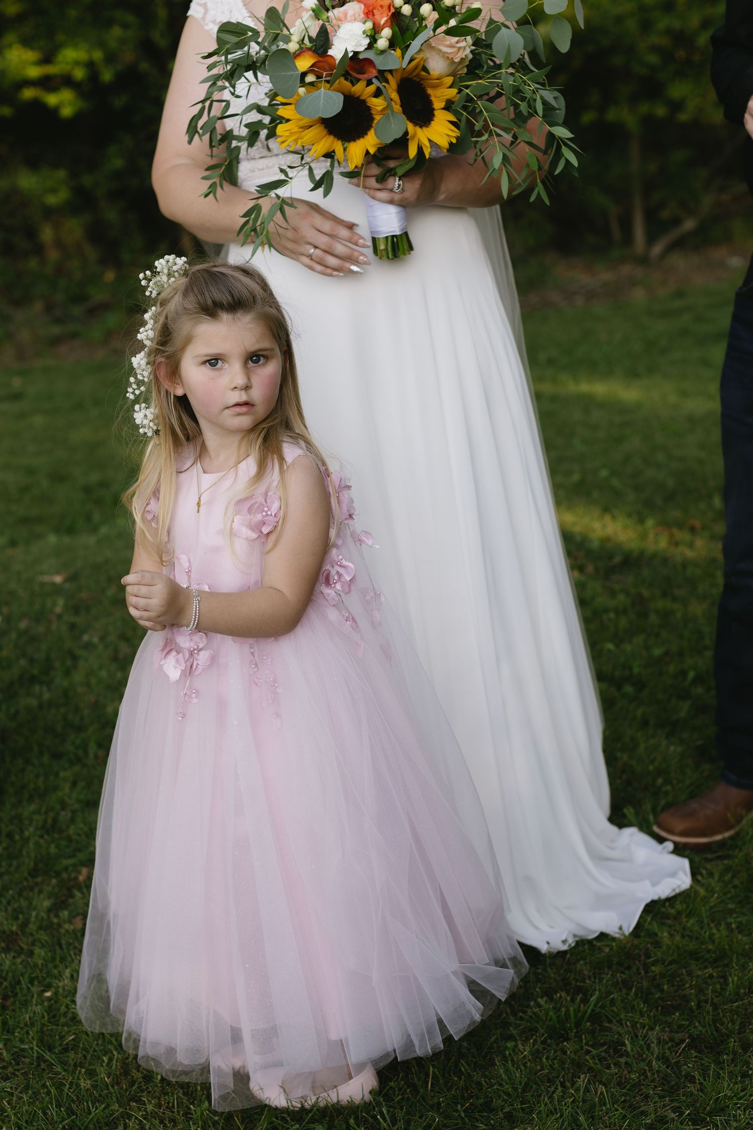 A flower girl in a pink tulle dress stands next to a white wedding gown holding a sunflower bouquet.