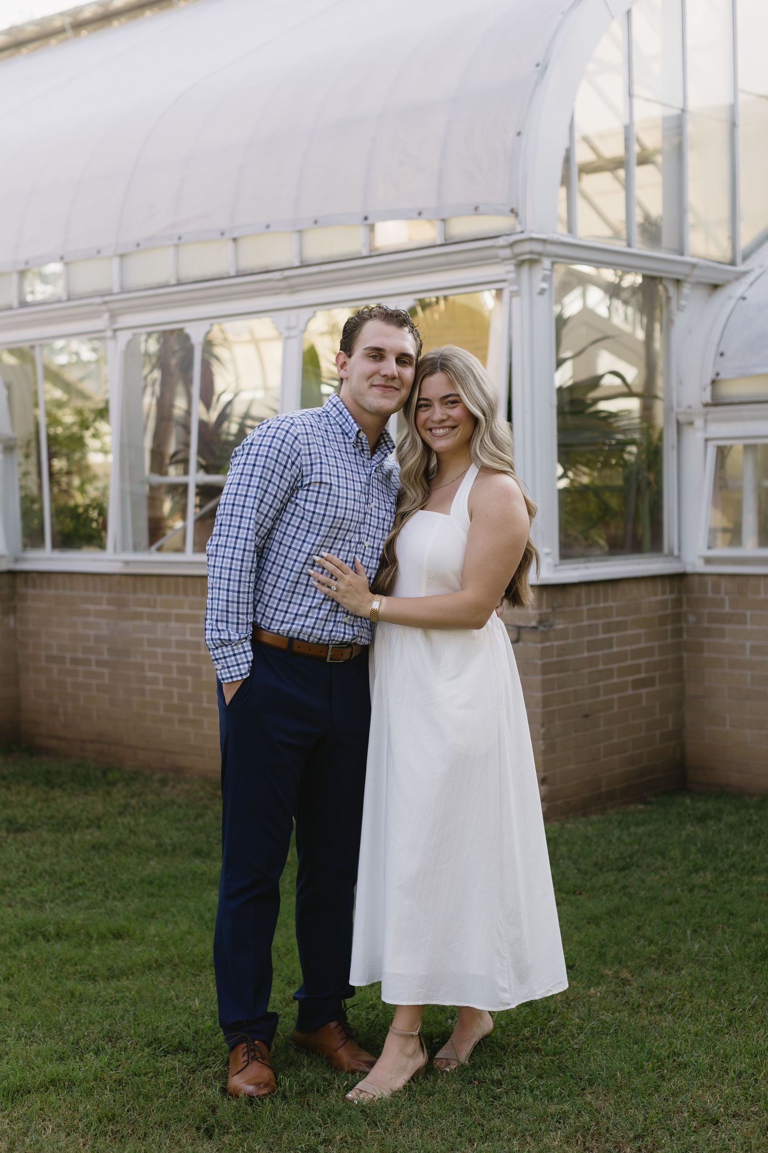 A couple poses together on a grassy lawn near a conservatory in casual summer attire.