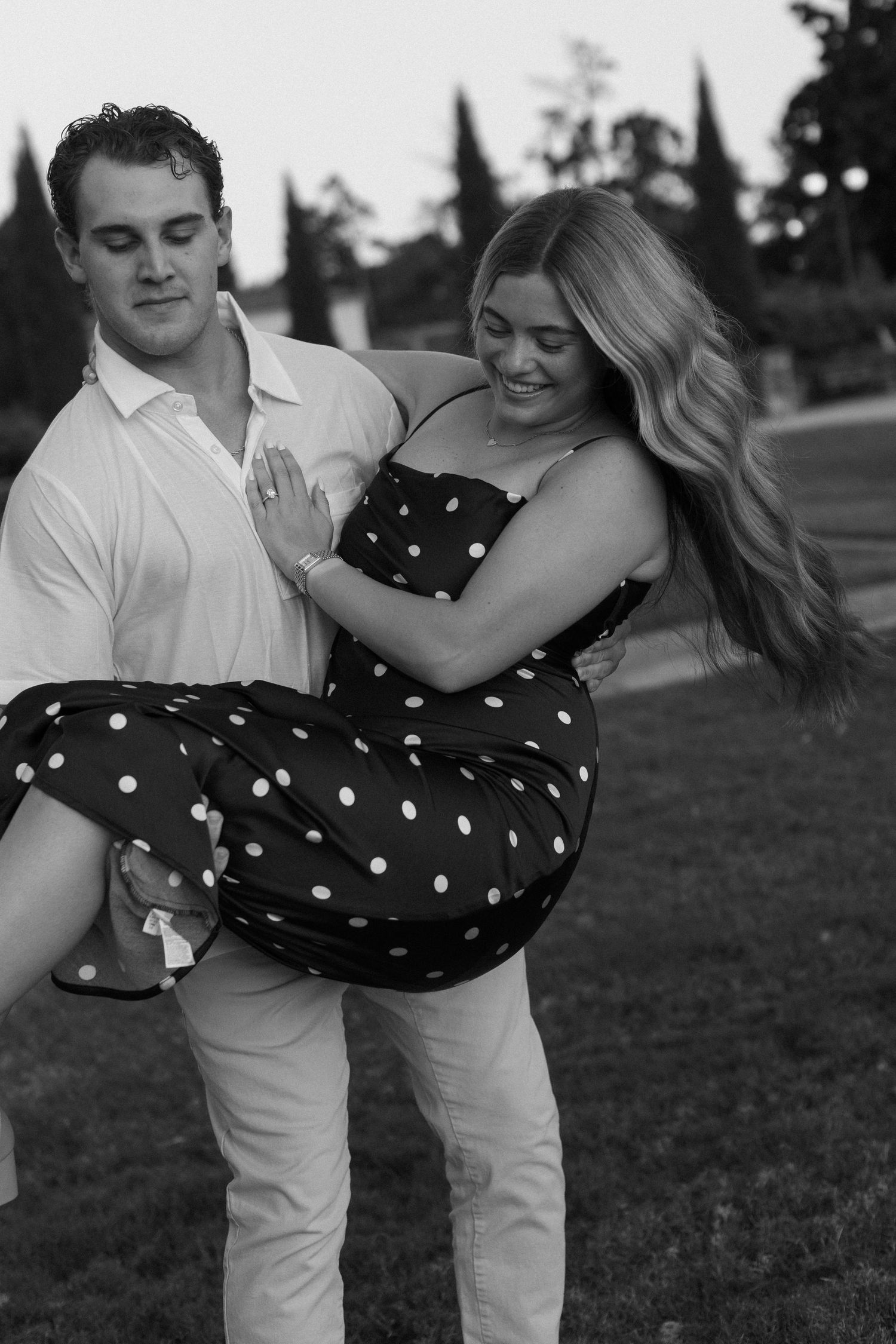 A couple in a polka dot dress and white shirt dance together playfully in a grass field at sunset.